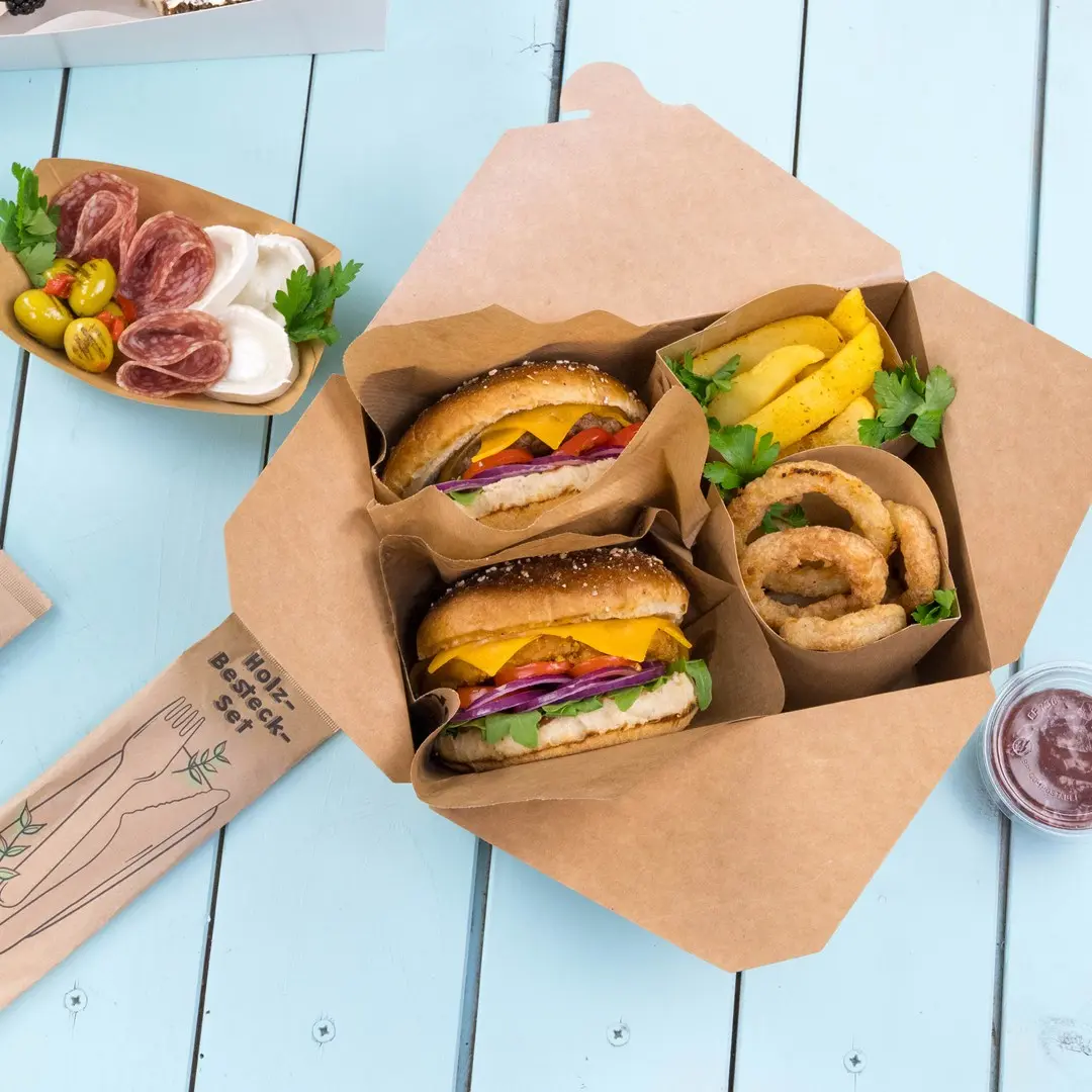Boîte à emporter contenant deux hamburgers, des rondelles d'oignon, des quartiers de pommes de terre et un petit contenant de sauce sur une surface en bois bleu.