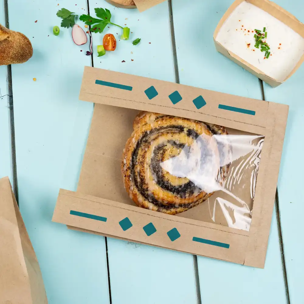 Sac en papier brun avec une fenêtre transparente présentant une pâtisserie en spirale sur une table bleue.