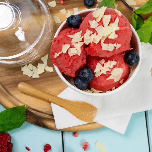 Bol de sorbet framboise garni de myrtilles et copeaux de chocolat blanc, cuillère en bois et feuilles de menthe sur une surface en bois.