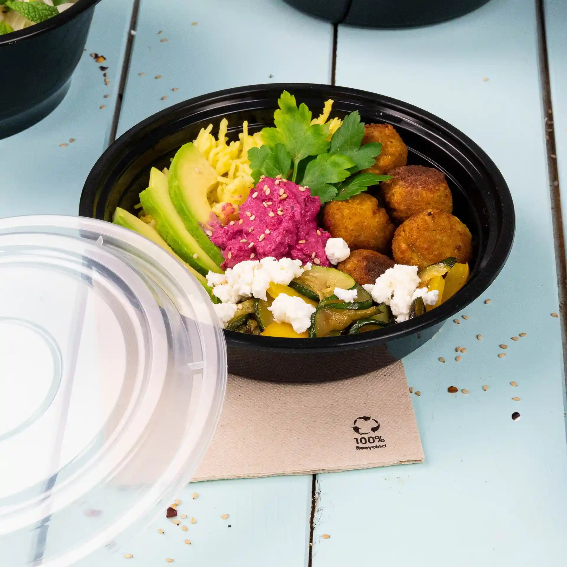 Assiette ronde réutilisable noire avec divers aliments, y compris de l'avocat, des falafels et une sauce rose, partiellement couverte par un couvercle transparent.