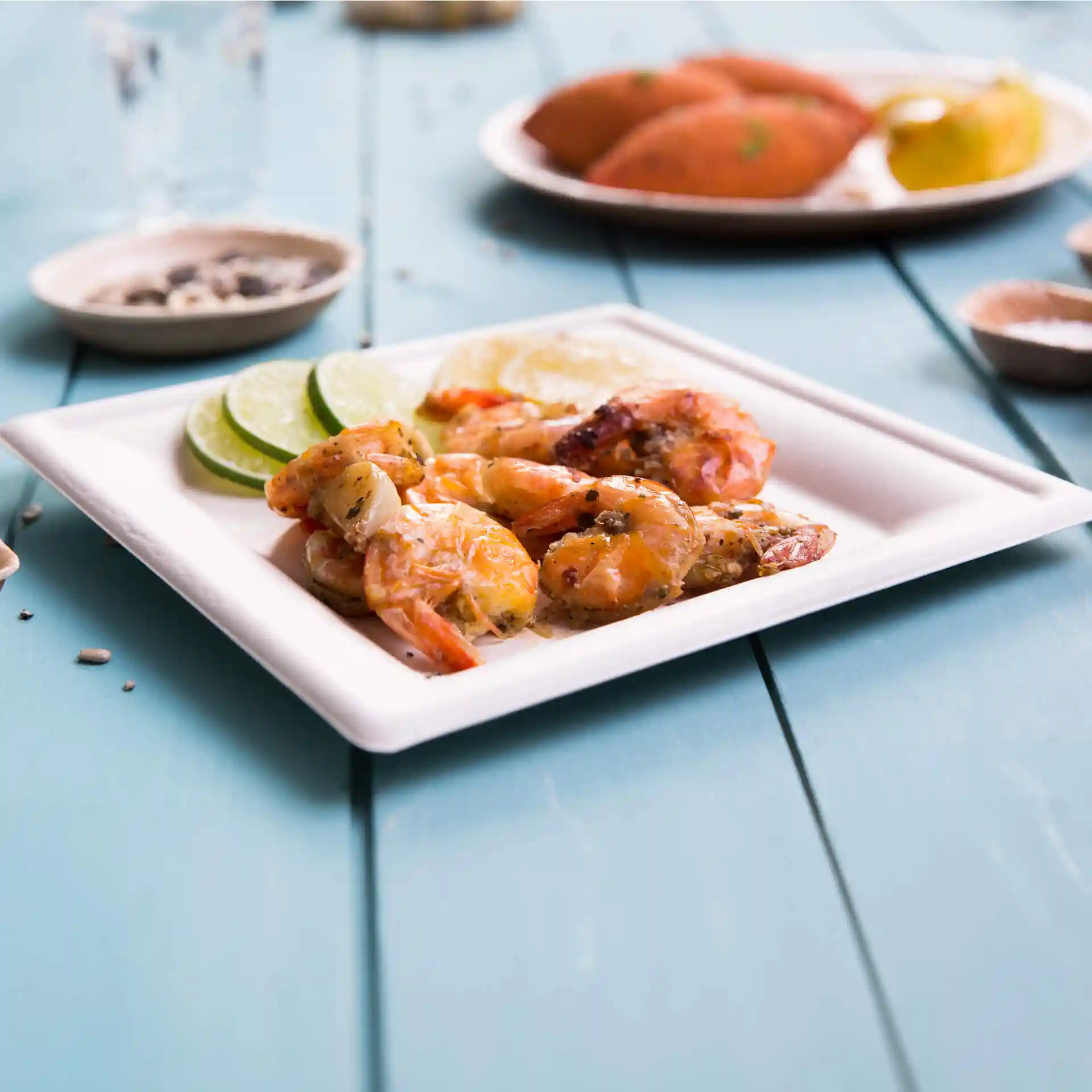 Assiette carrée blanche en canne à sucre, contenant des crevettes grillées et des tranches de citron vert, sur une table en bois bleu.