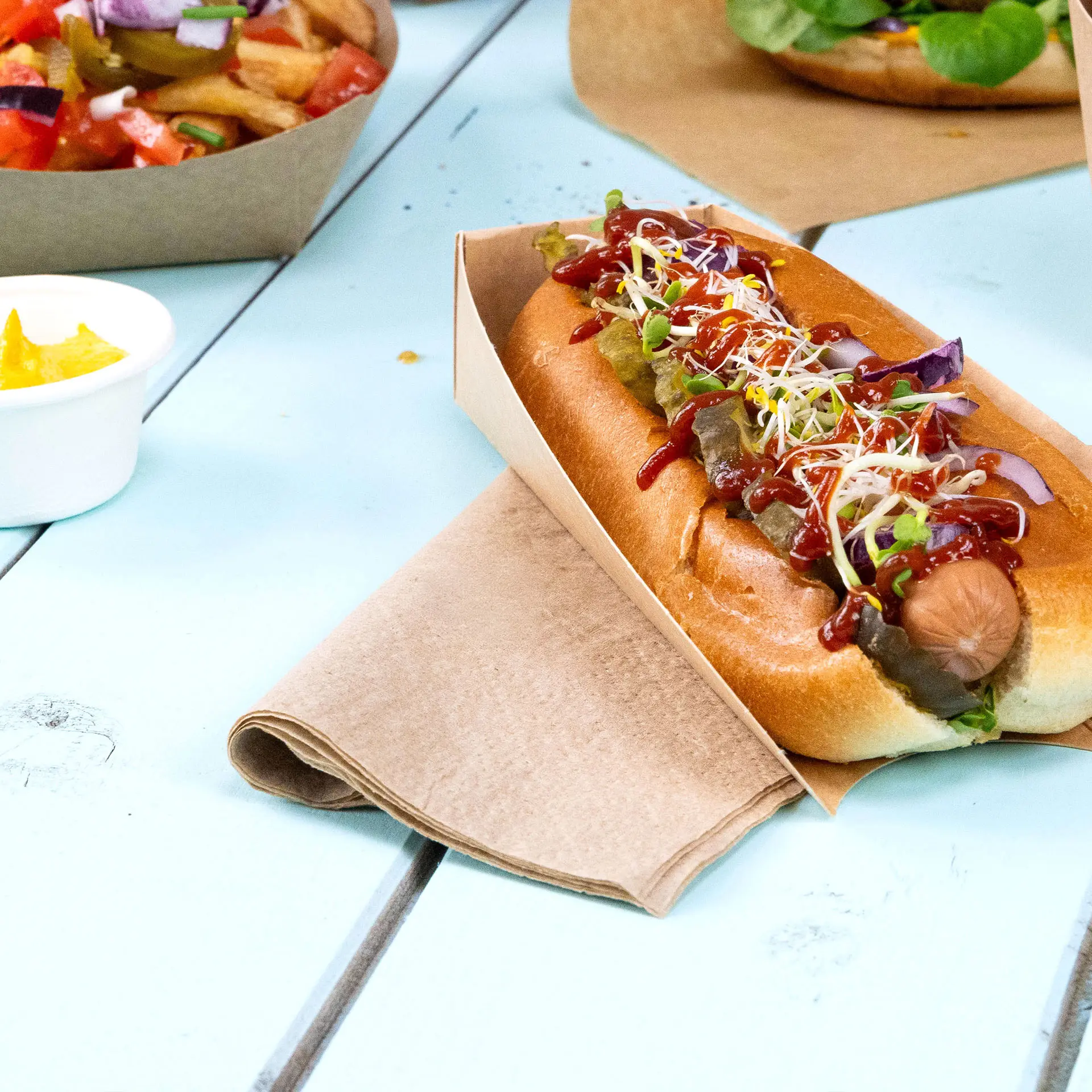 Hot-dog dans un support en papier, garni de ketchup, moutarde, cornichons et pousses, avec une serviette carrée non blanchie à côté.