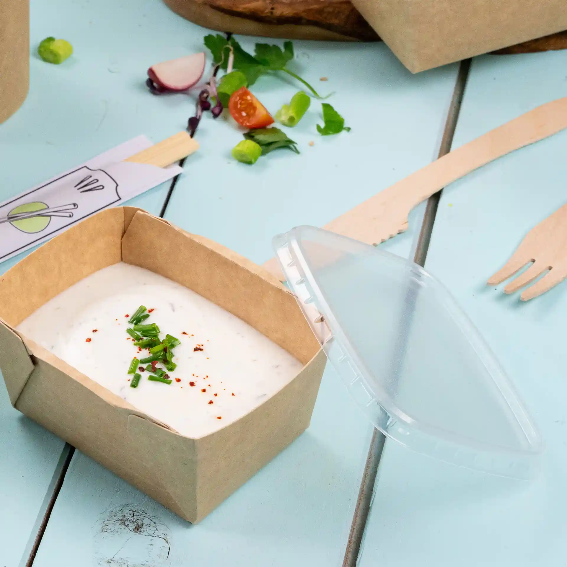 Conteneur en carton rectangulaire avec un couvercle en rPET transparent, contenant une sauce crémeuse garnie de ciboulette hachée et de flocons de piment rouge.