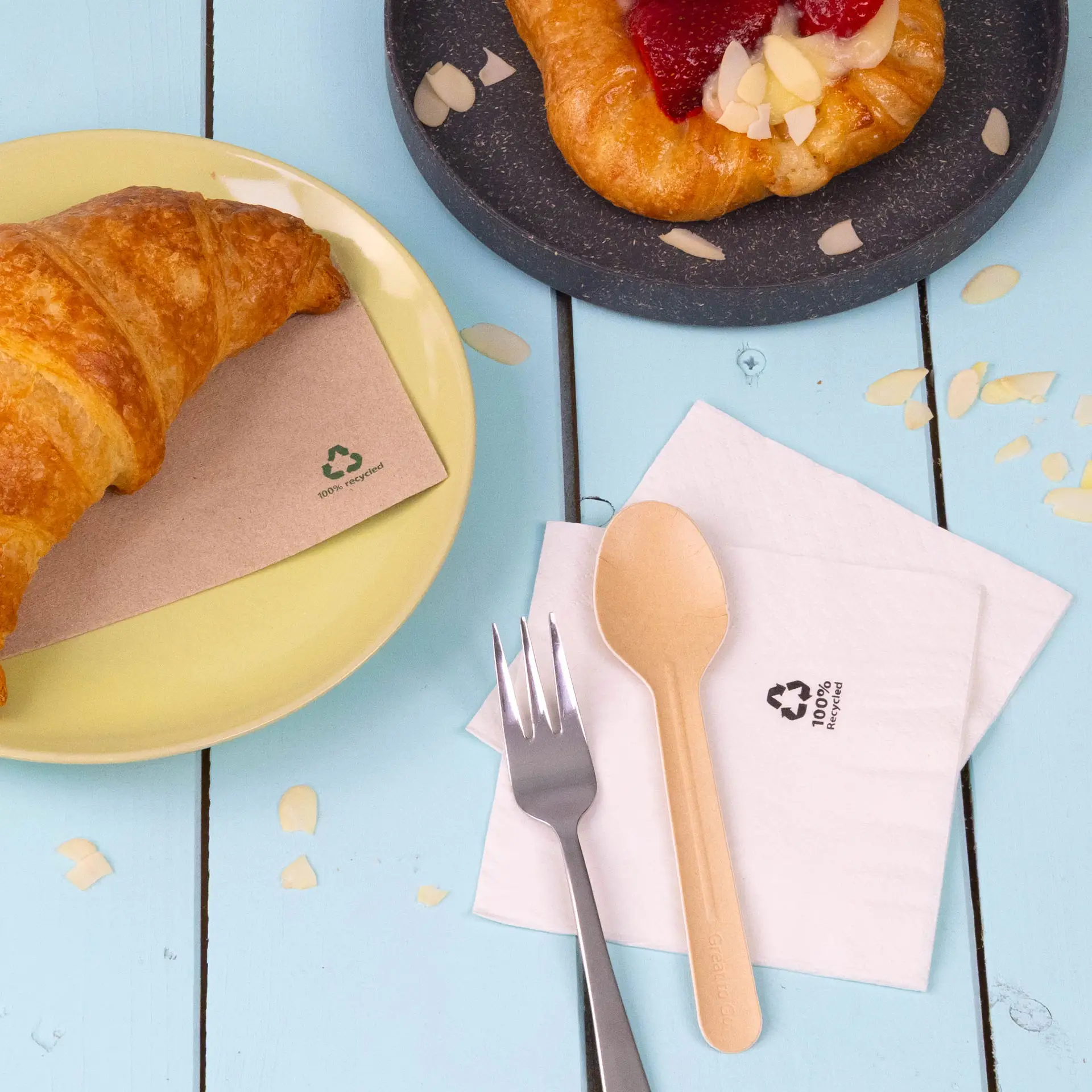 Un croissant sur une assiette jaune à côté d'une serviette en papier recyclé 2 plis blanche et d'une cuillère et d'une fourchette en bois écologiques.