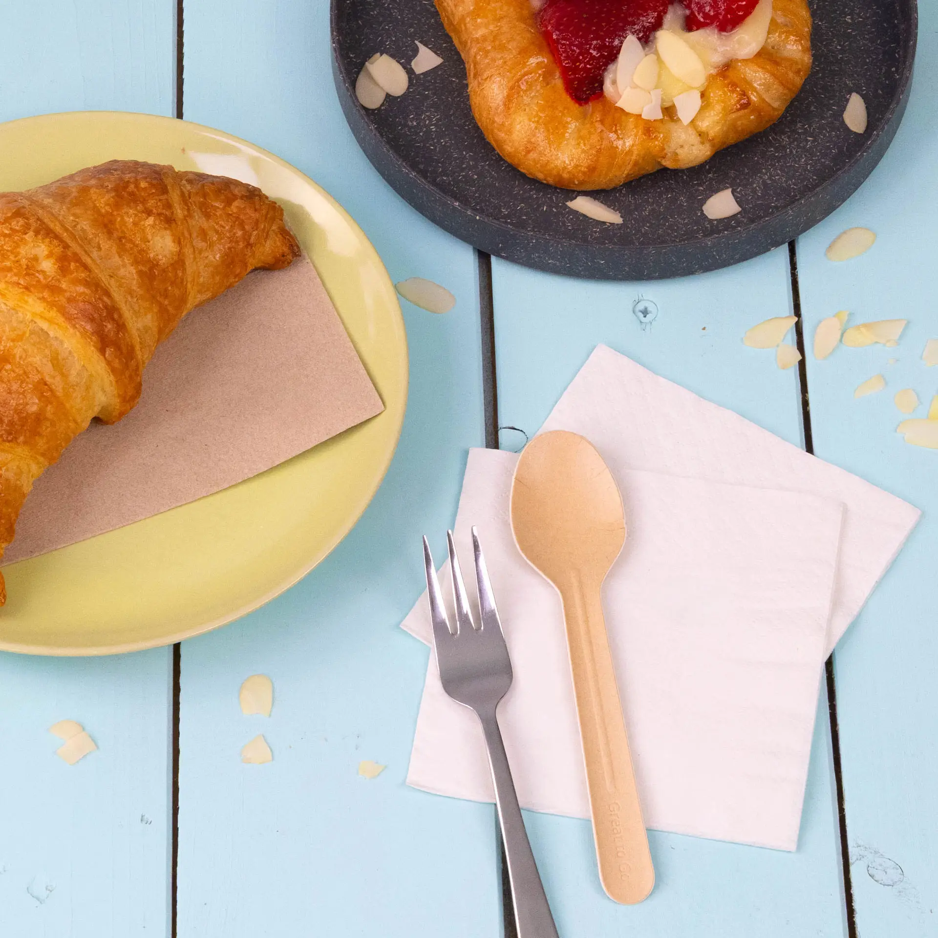 Image d'un croissant sur une assiette jaune, une pâtisserie sur une assiette sombre, une cuillère en bois, une fourchette en argent et des serviettes blanches.