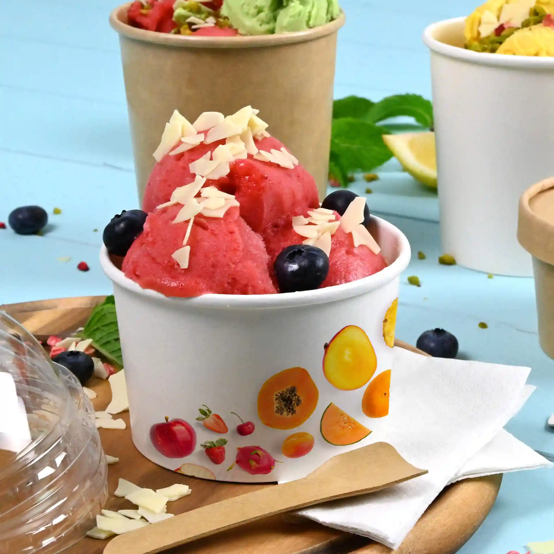 Coupe ronde blanche de glace remplie de sorbet rose, garnie d'amandes effilées et de myrtilles, sur un plateau en bois.