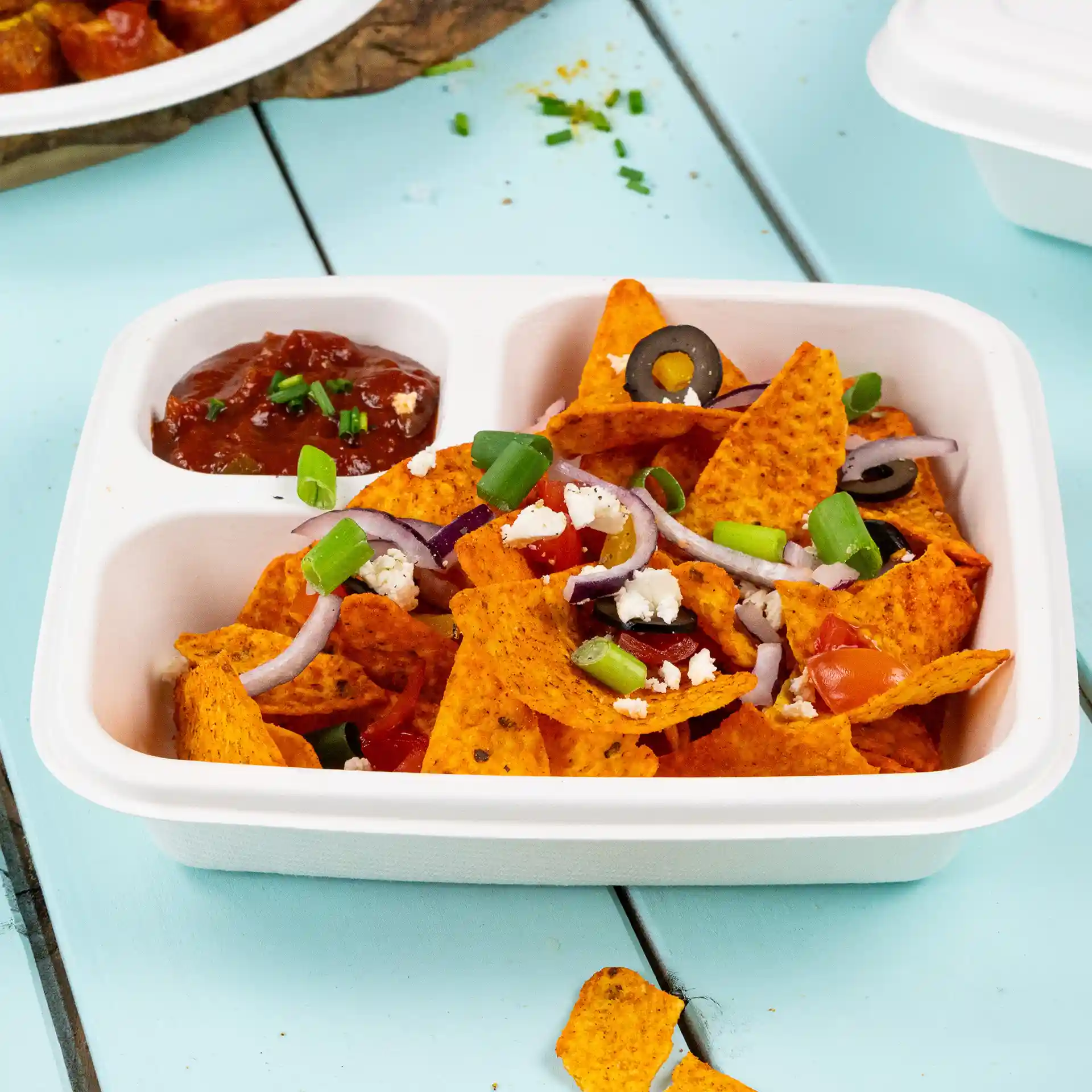 Assiette rectangulaire blanche en canne à sucre remplie de nachos, garnie de tomates, olives, oignons et oignons verts, avec salsa à côté.