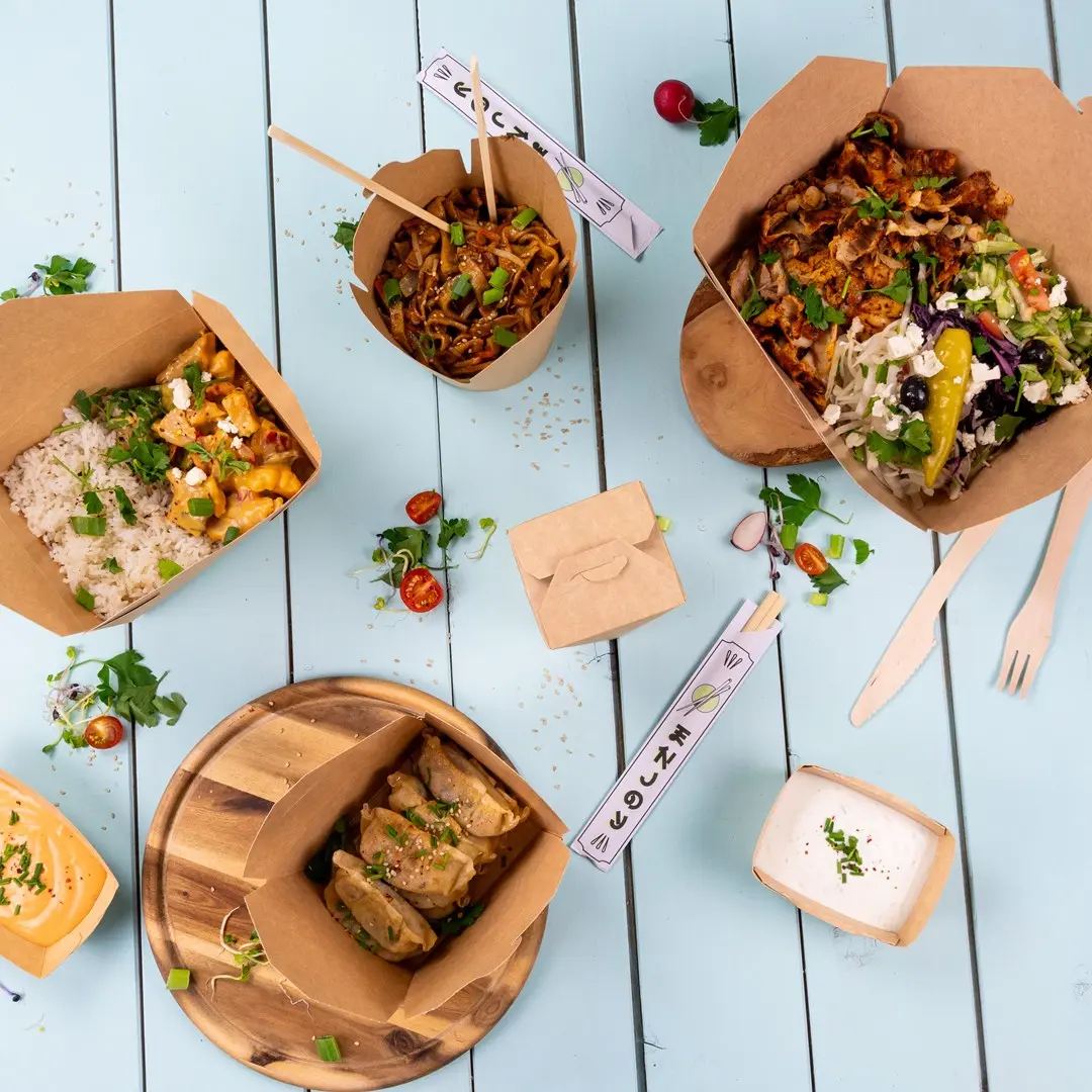 Contenants de nourriture à emporter assortis avec des nouilles, du riz, de la salade et des raviolis sur une surface en bois bleu.