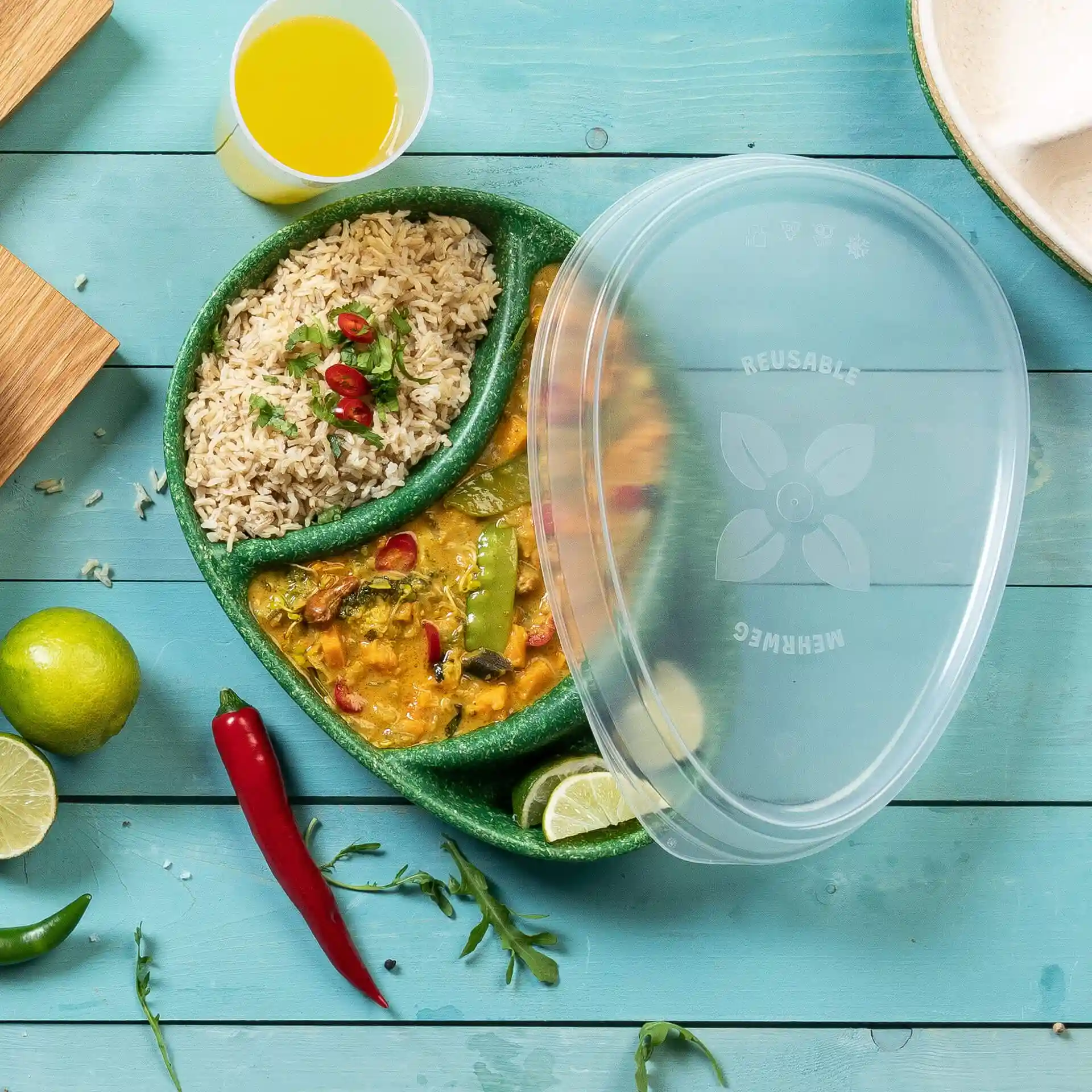 Couvercle réutilisable transparent sur une assiette divisée contenant du riz et du curry, avec du citron vert et des piments sur une surface en bois bleu.
