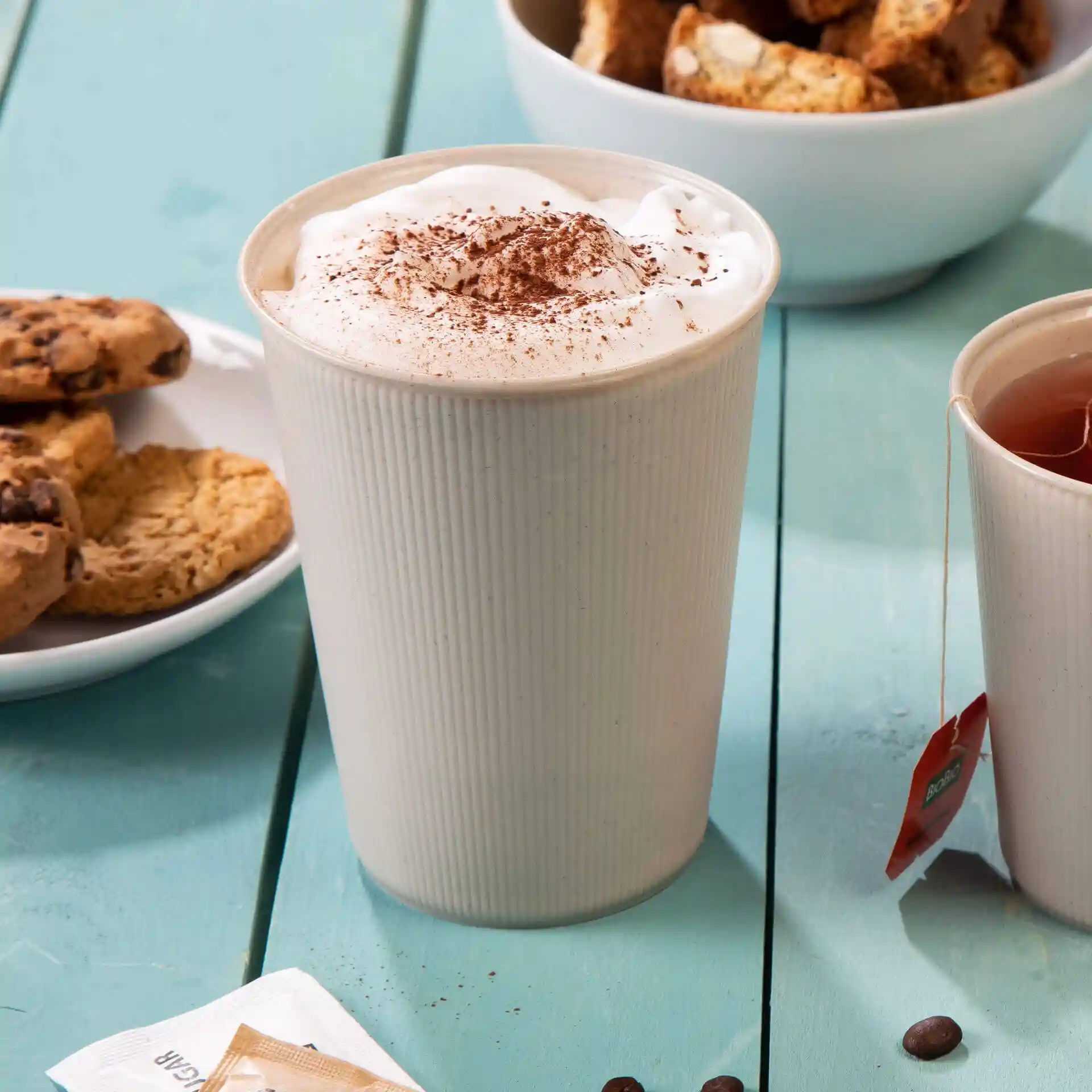 Gobelet réutilisable à rayures blanc crème rempli de crème fouettée et de cacao, avec des biscuits et du thé à proximité sur une table bleue.