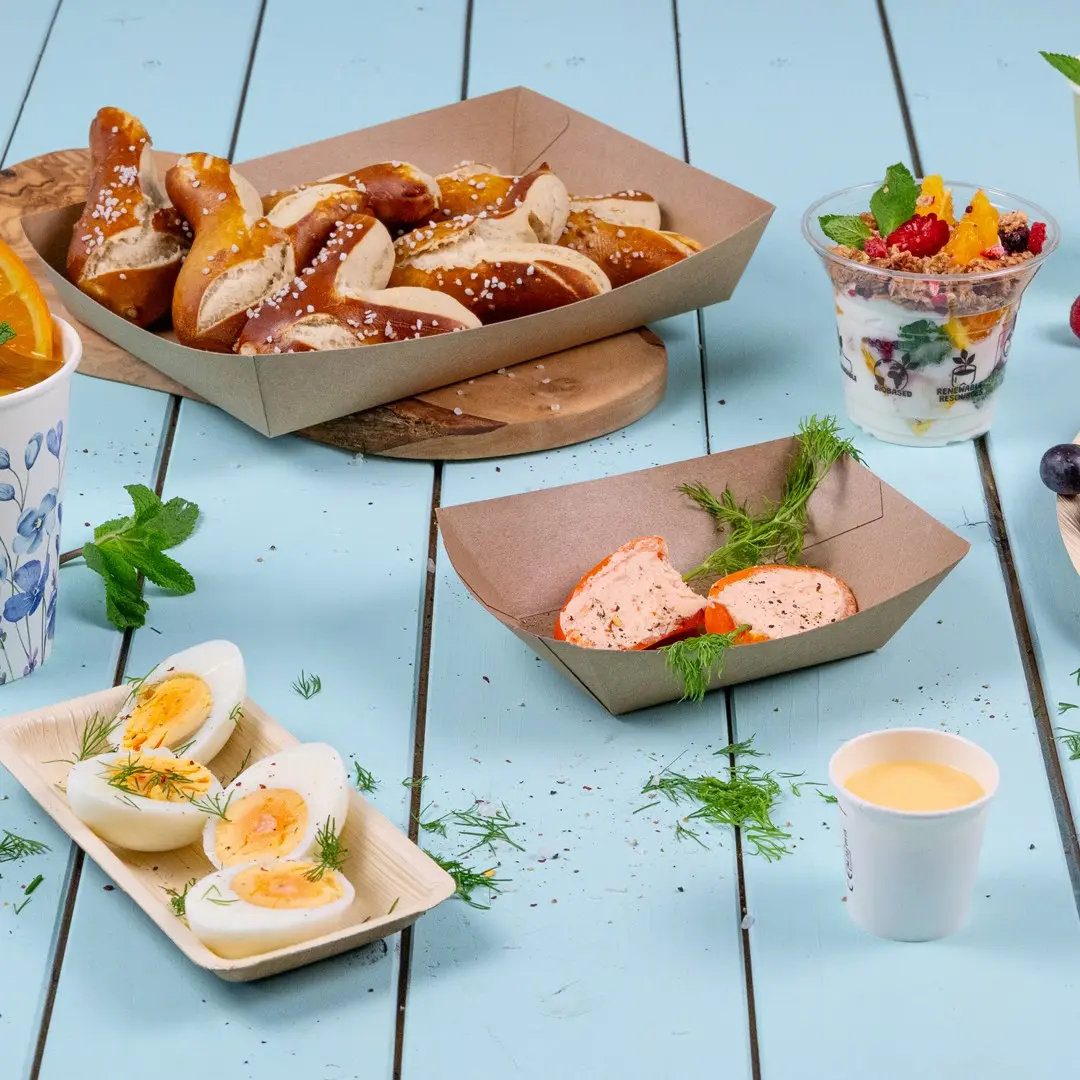 Assortiment de snacks comprenant des bretzels, des œufs durs, un parfait au yaourt et un petit pot de sauce sur une surface en bois bleu.