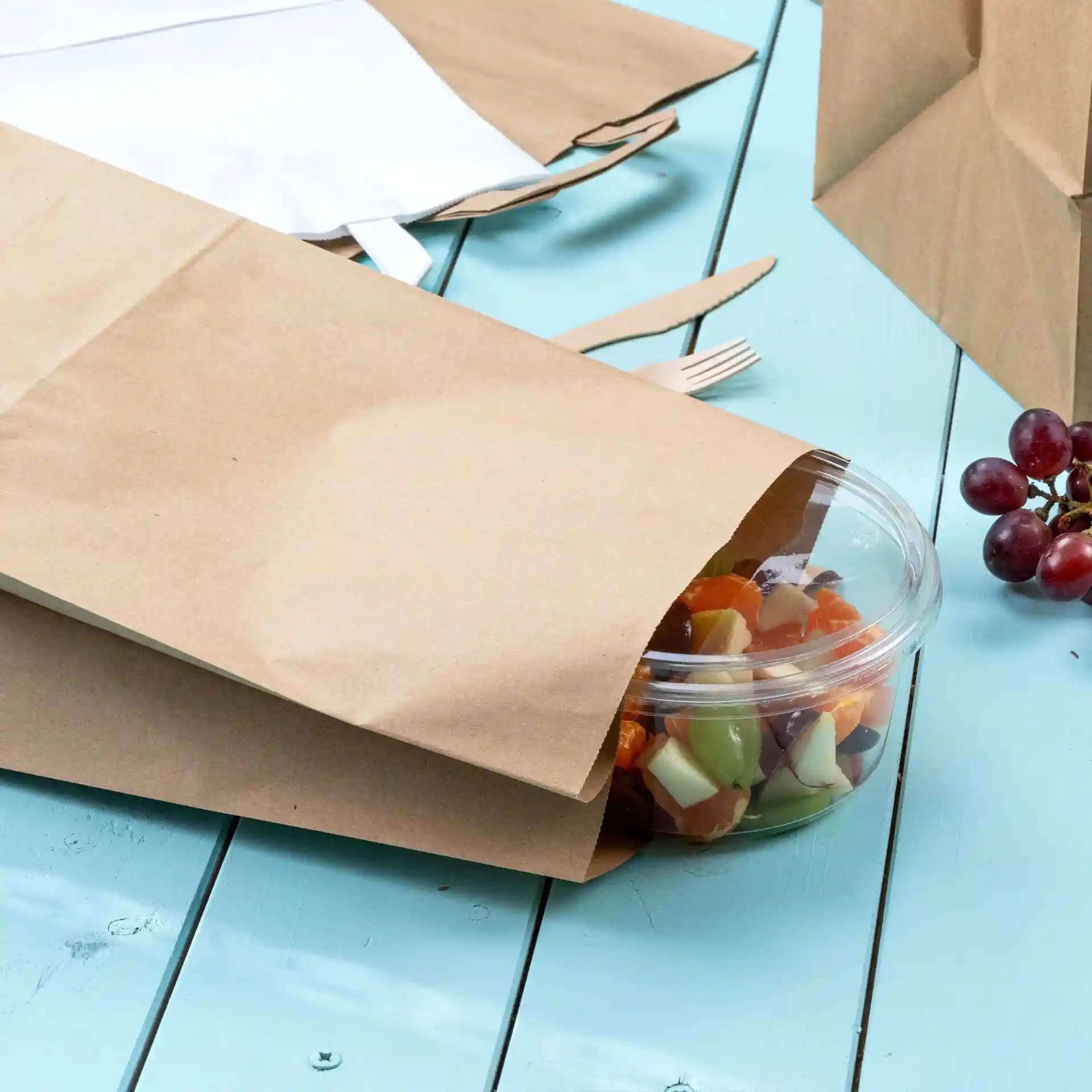 Sac de courses en papier kraft marron couvrant partiellement un contenant en plastique transparent avec des fruits mélangés, sur une surface en bois bleu.
