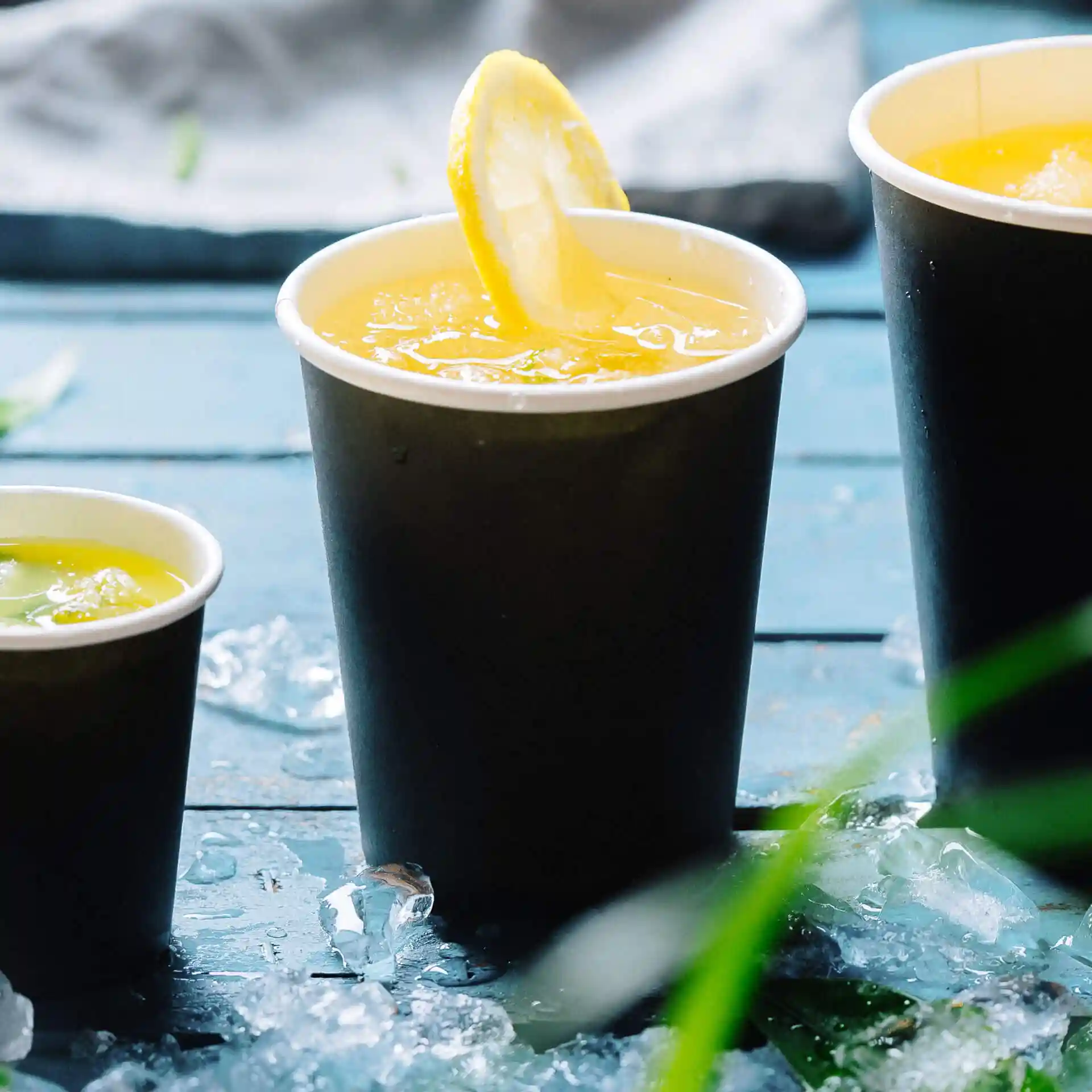 Trois gobelets en papier noirs remplis de boissons, l'un surmonté d'une tranche de citron, posés sur une surface en bois bleu avec de la glace.