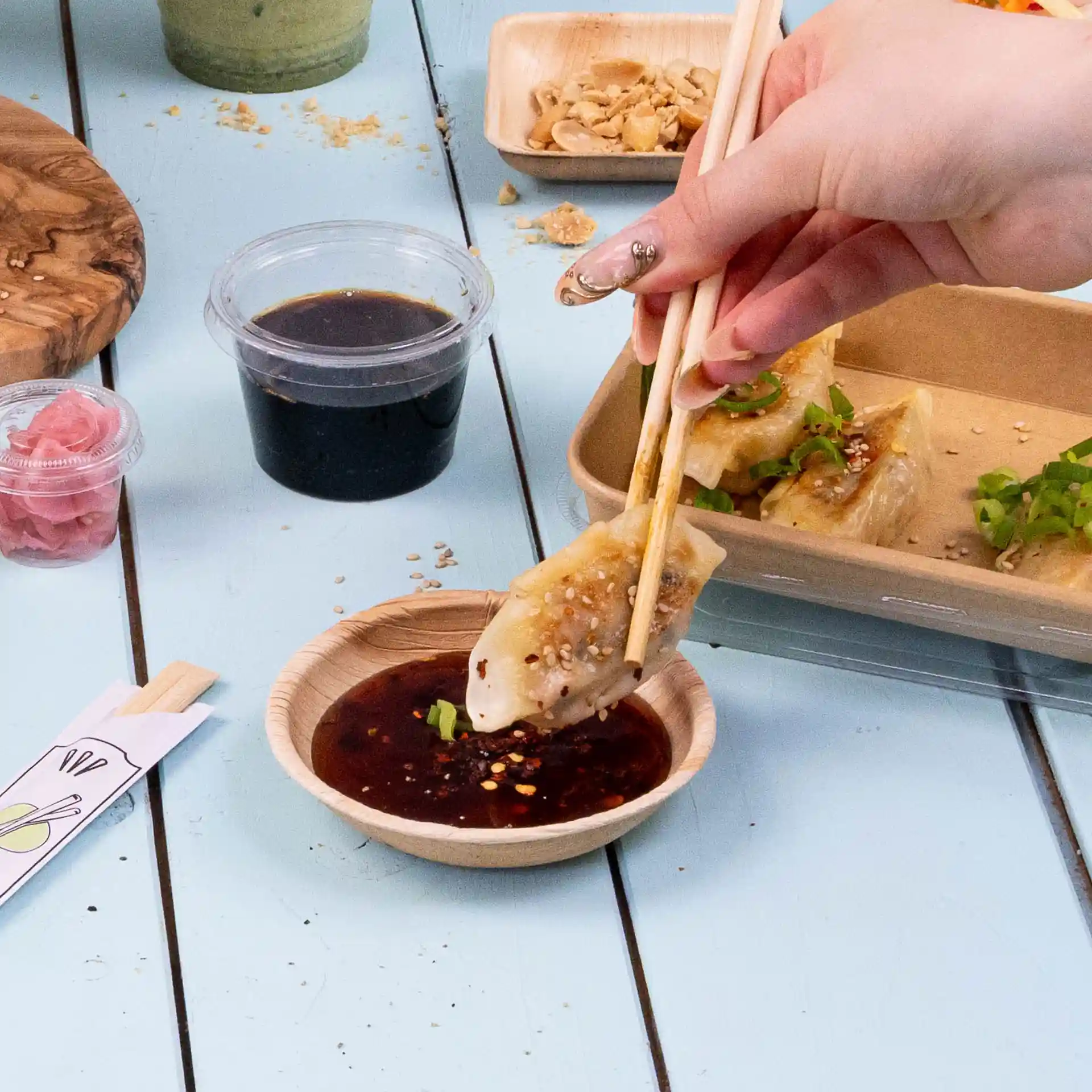 Une main tenant des baguettes plonge un dumpling dans un bol rond en feuille de palmier rempli de sauce sombre.