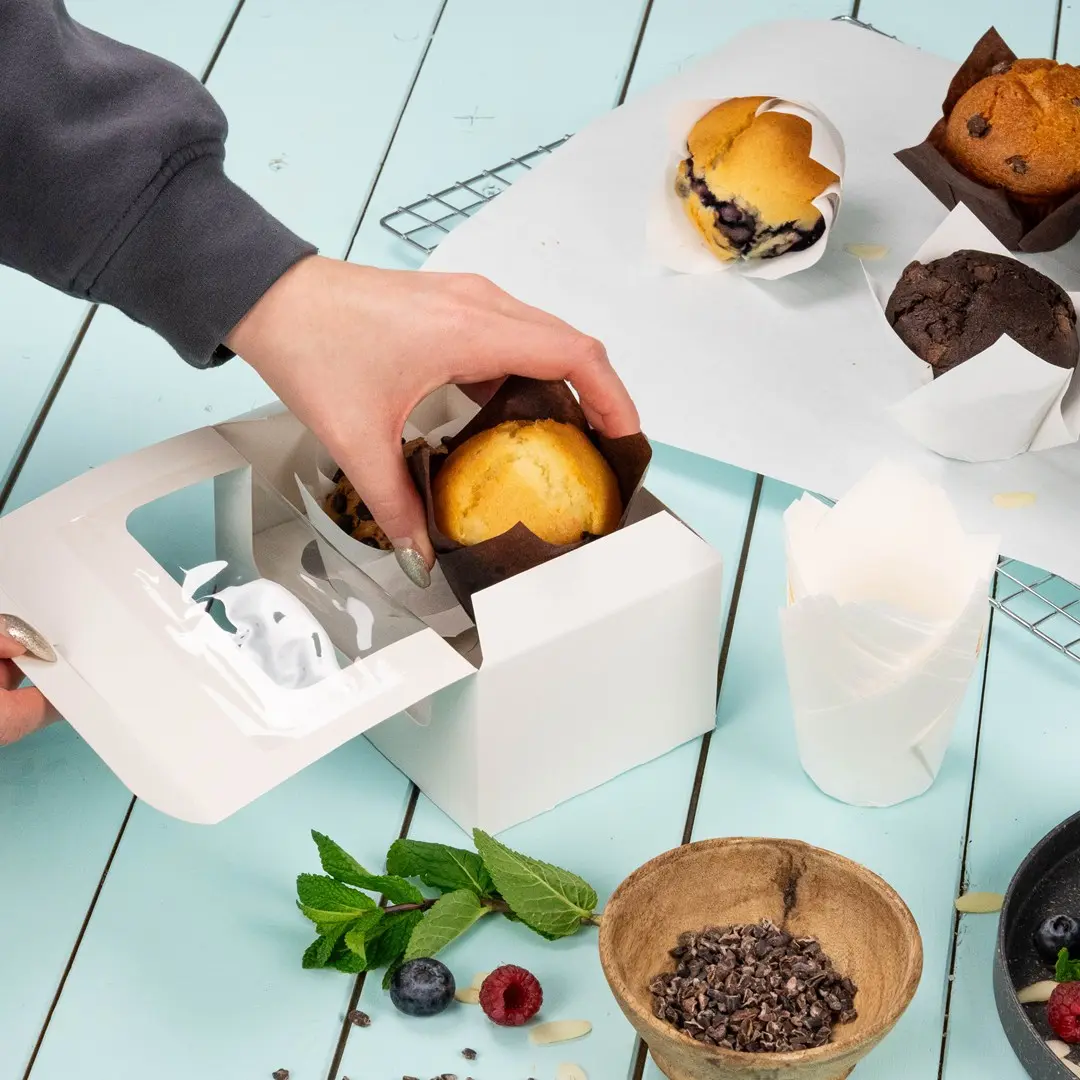 Main plaçant un muffin dans une boîte à cupcakes blanche avec plusieurs produits de boulangerie et ingrédients sur une surface bleue.