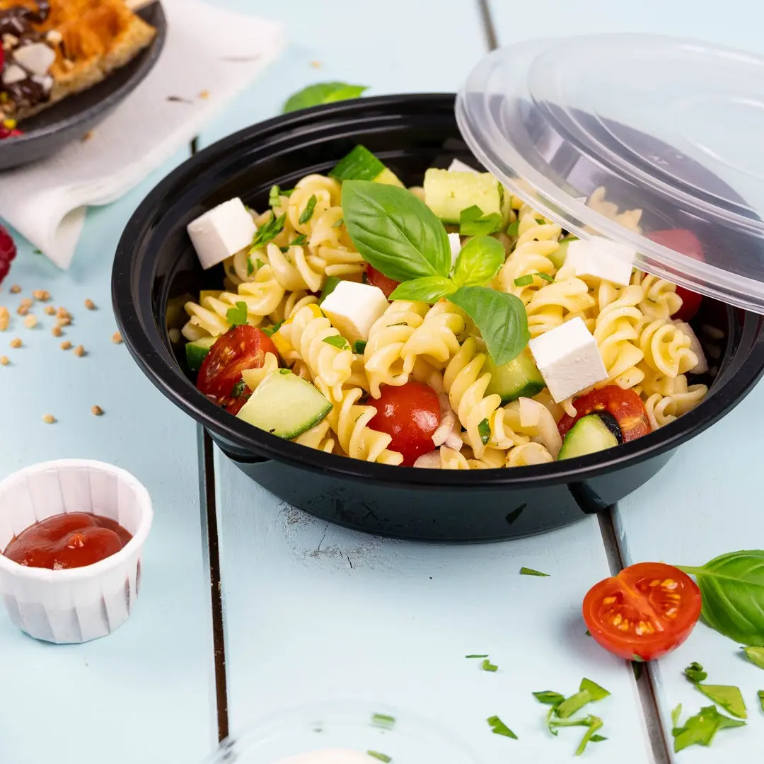 Bol noir avec salade de pâtes contenant des rotinis, tomates cerises, concombre, feta et basilic, avec un côté de ketchup.