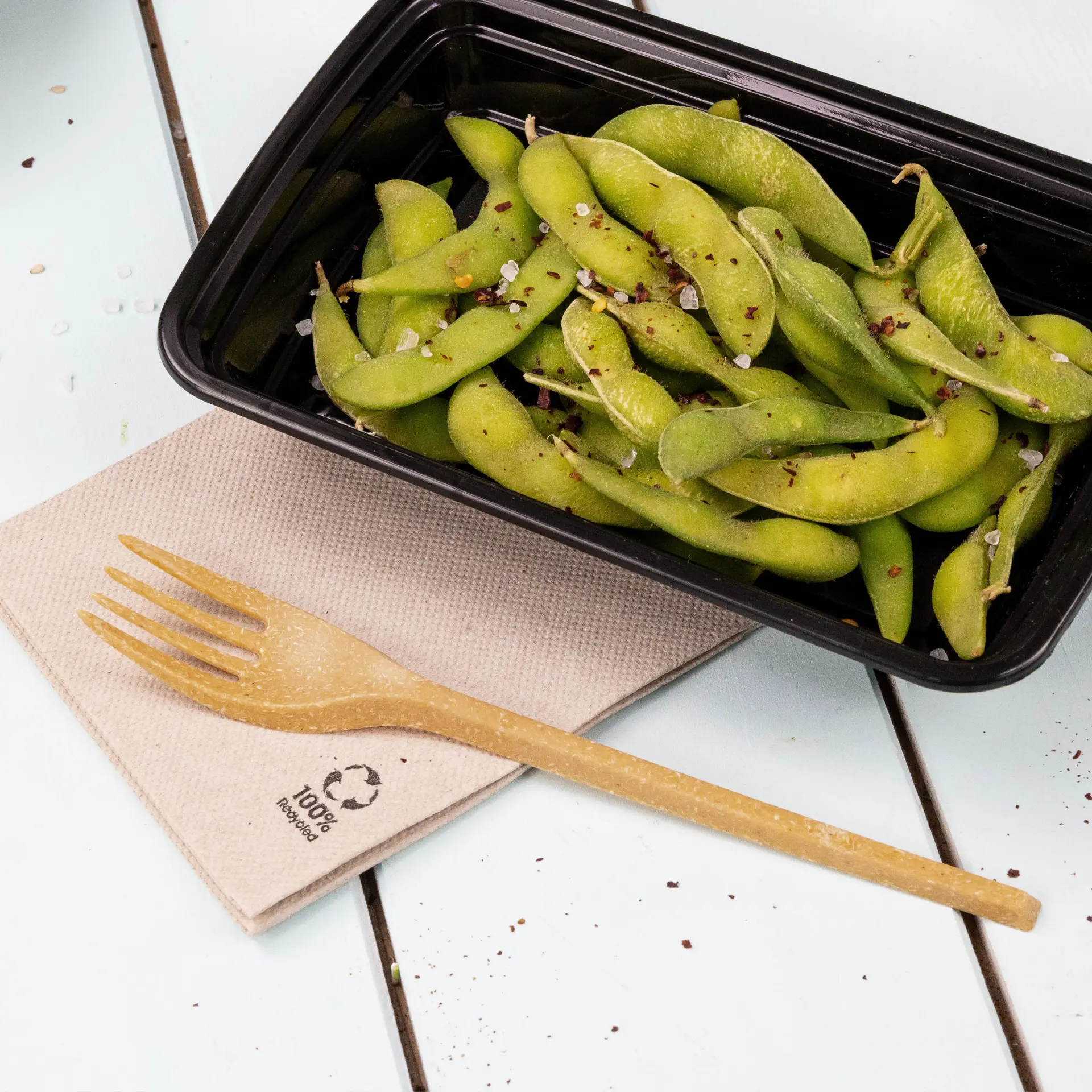 Un conteneur noir rempli d'edamame verts garni d'épices, accompagné d'une fourchette biodégradable sur une serviette en papier recyclé.