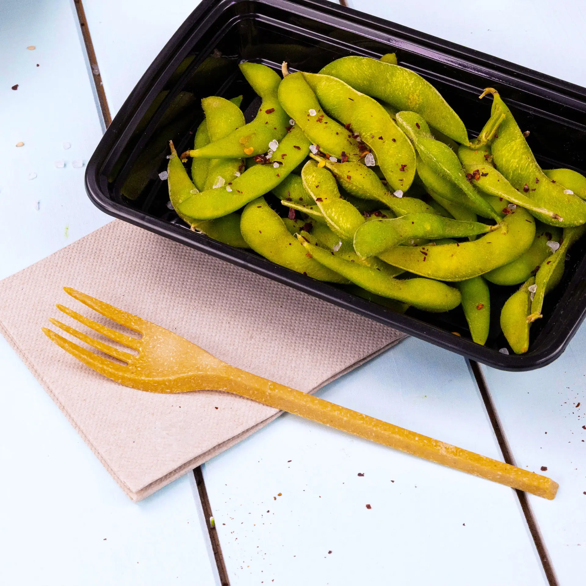 Image d'un conteneur noir rempli de fèves edamame vertes, une fourchette jaune sur une serviette beige à côté.