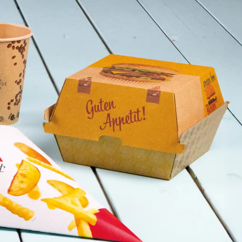 Boîte de fast-food avec illustration de burger et texte "Bon appétit!"; gobelet en papier et emballage de frites à proximité sur une surface en bois.
