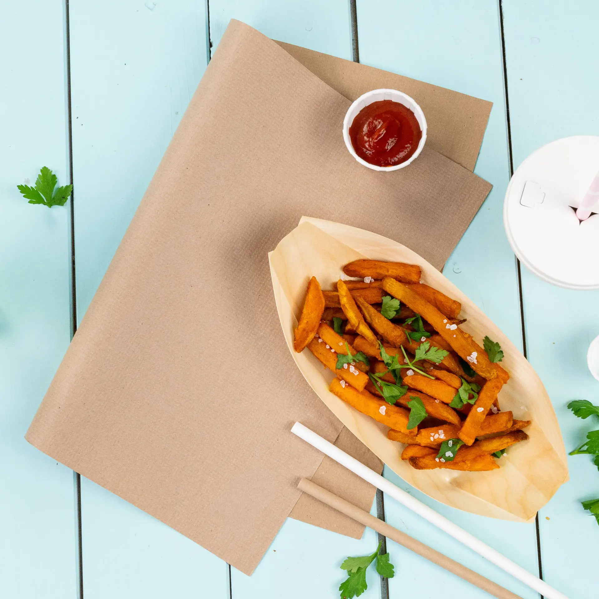 Nappe en papier brun avec des frites de patate douce dans un récipient en forme de bateau, ketchup et coriandre sur fond bleu.