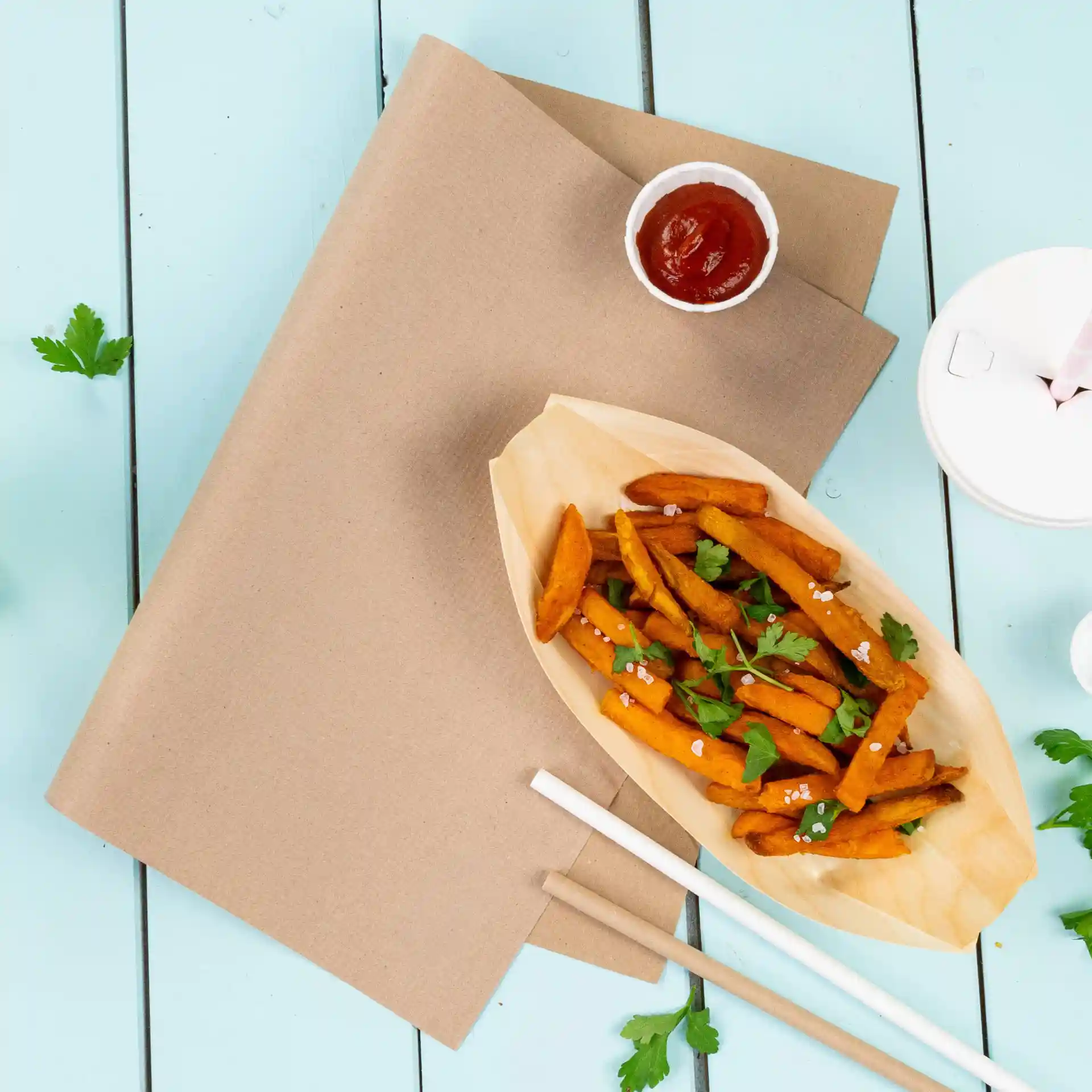 Nappe en papier brun avec des frites de patate douce dans un récipient en forme de bateau, ketchup et coriandre sur fond bleu.