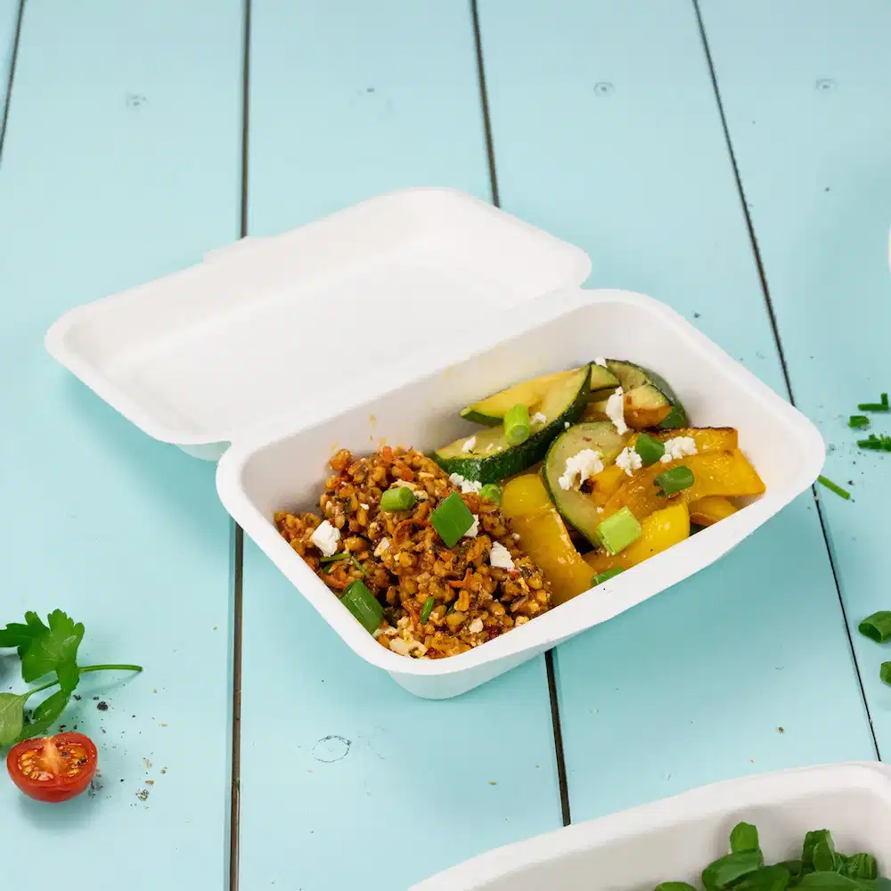 Conteneur alimentaire blanc avec un mélange de grains cuits, courgettes, poivrons, garni d'oignons verts et de fromage émietté.