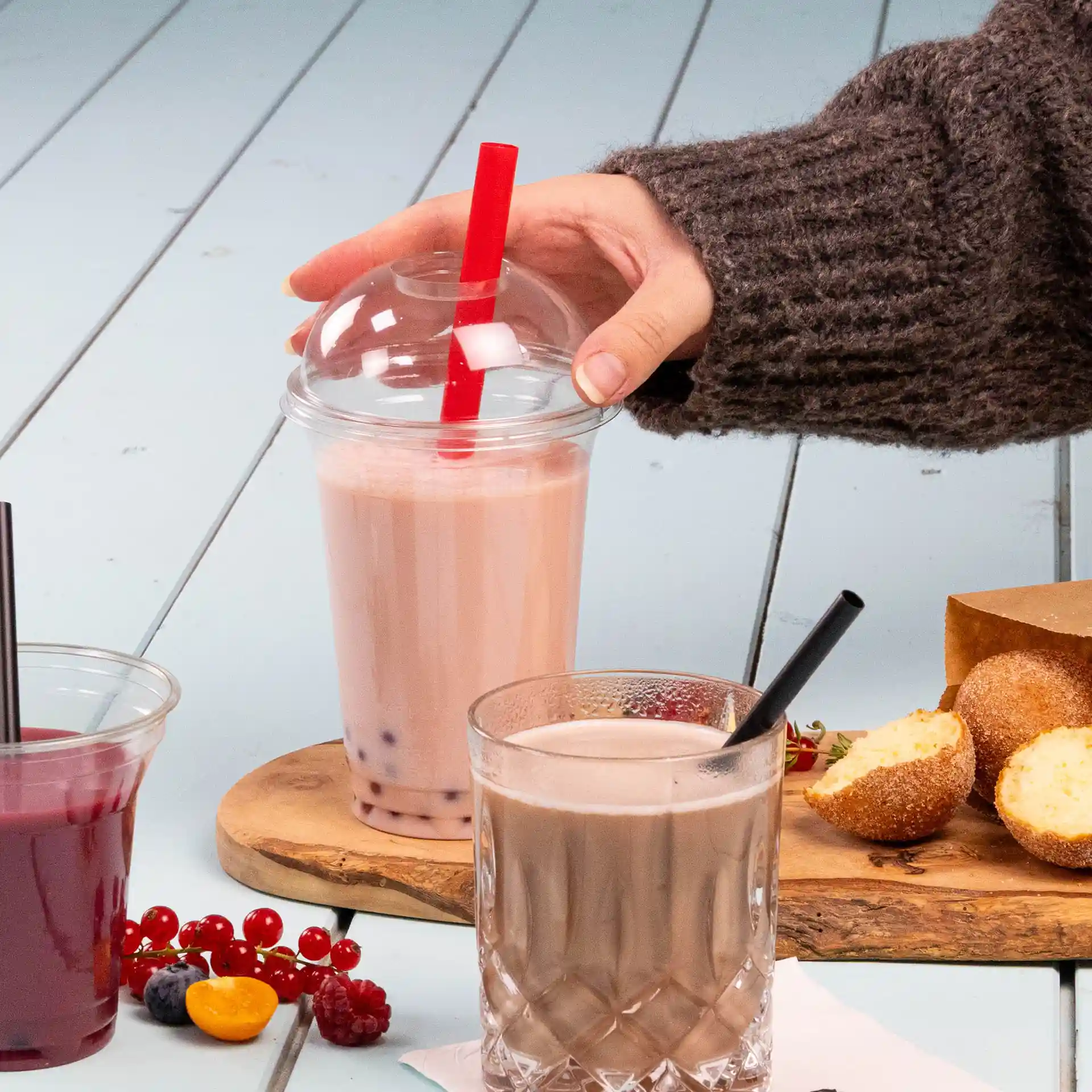 Image d'une main tenant un verre de bubble tea avec une paille rouge, à côté d'un verre de chocolat et de collations sur une planche en bois.