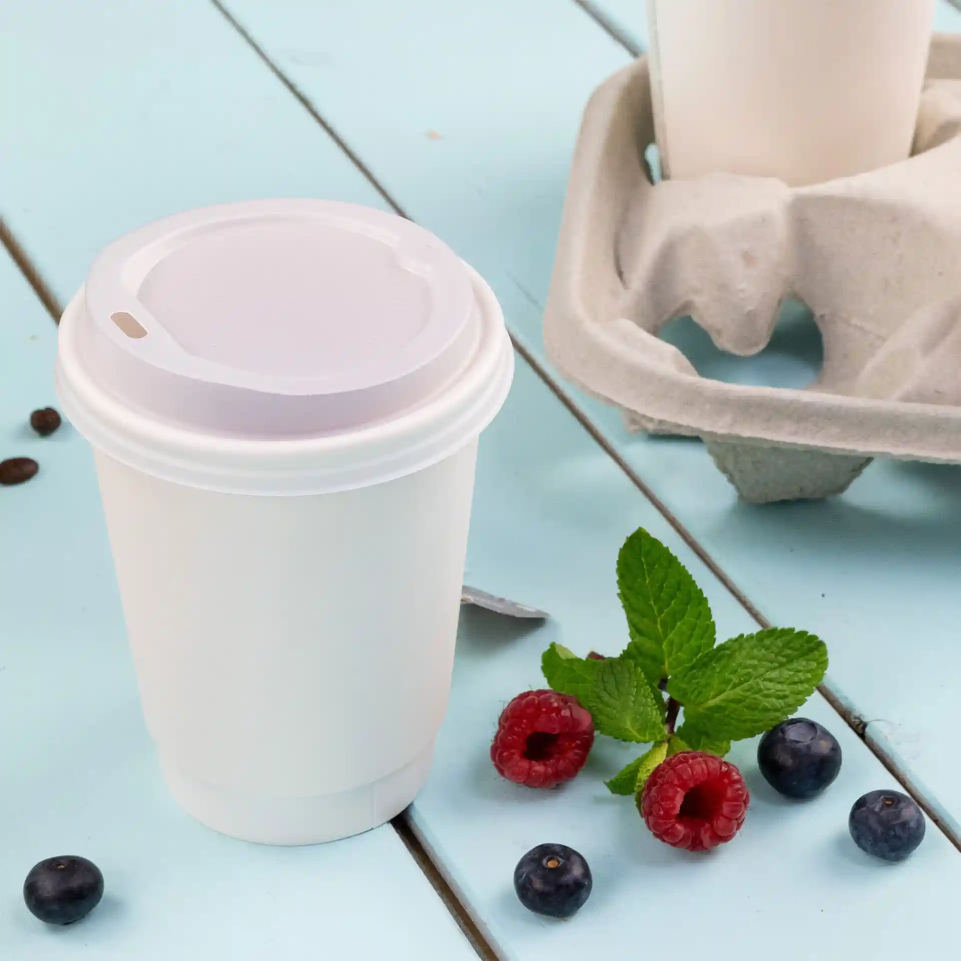 Tasse à café blanche à double paroi avec couvercle, 300 ml, placée sur une surface bleue avec des framboises, des myrtilles et des feuilles de menthe.