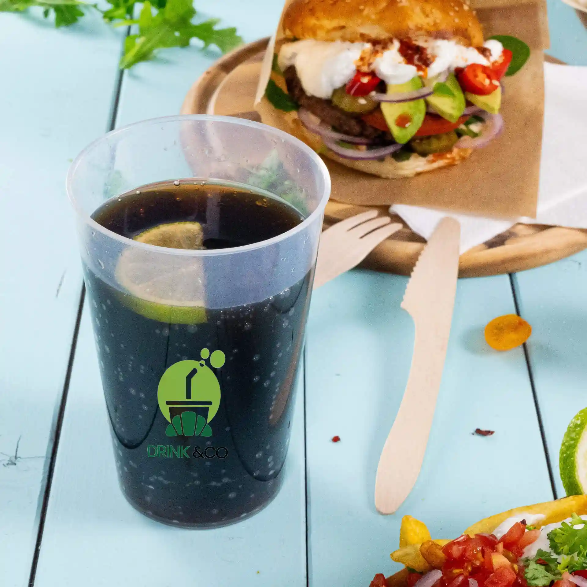 Gobelet réutilisable transparent avec logo, capacité de 500 ml, rempli de boisson sombre et d'une tranche de citron vert, placé à côté d'un burger et d'une salade.
