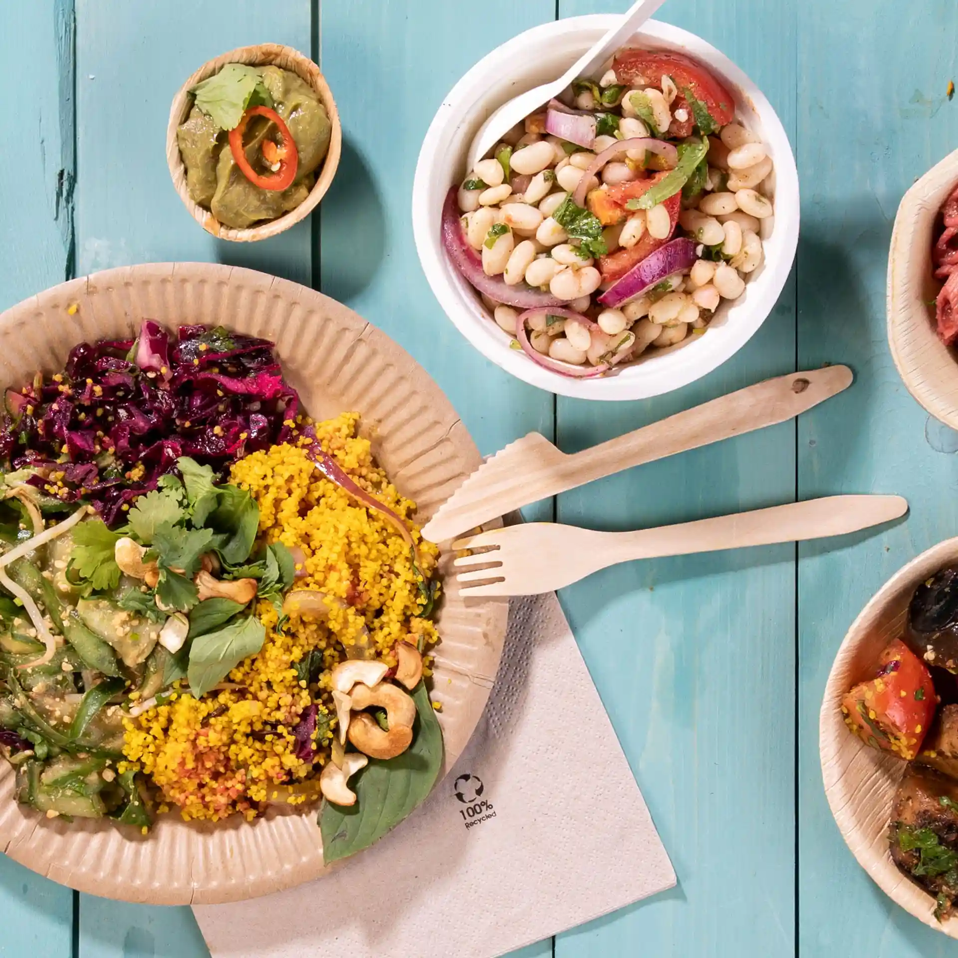 Image d'une assiette avec des aliments colorés, y compris du couscous, une salade et des garnitures, accompagnée de fourchettes en bois et de petits bols de garnitures.
