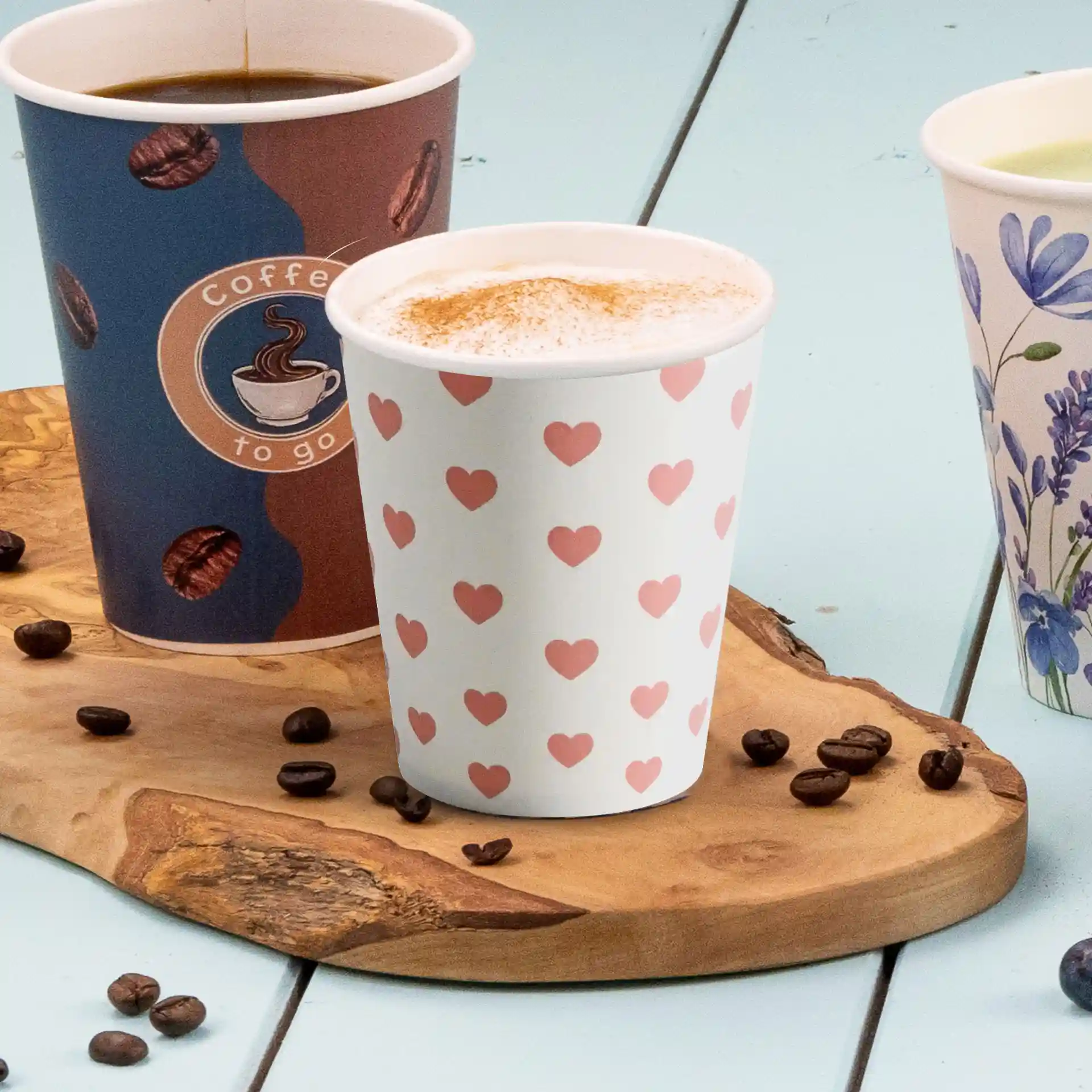 Trois gobelets en papier sur une planche en bois, un avec un motif en cœur, un avec un logo de café et un avec un design floral.