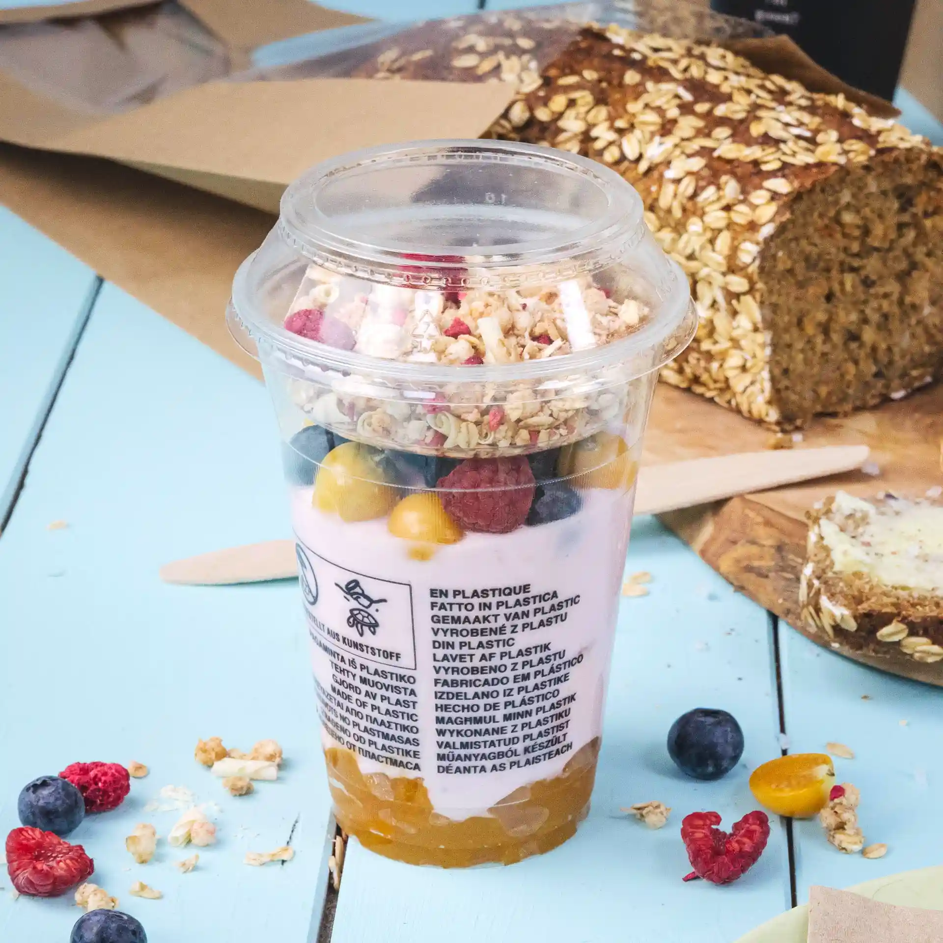 Gobelet smoothie PLA transparent de 12 oz rempli de yaourt, granola et fruits mélangés, posé sur une surface bleue avec du pain en arrière-plan.