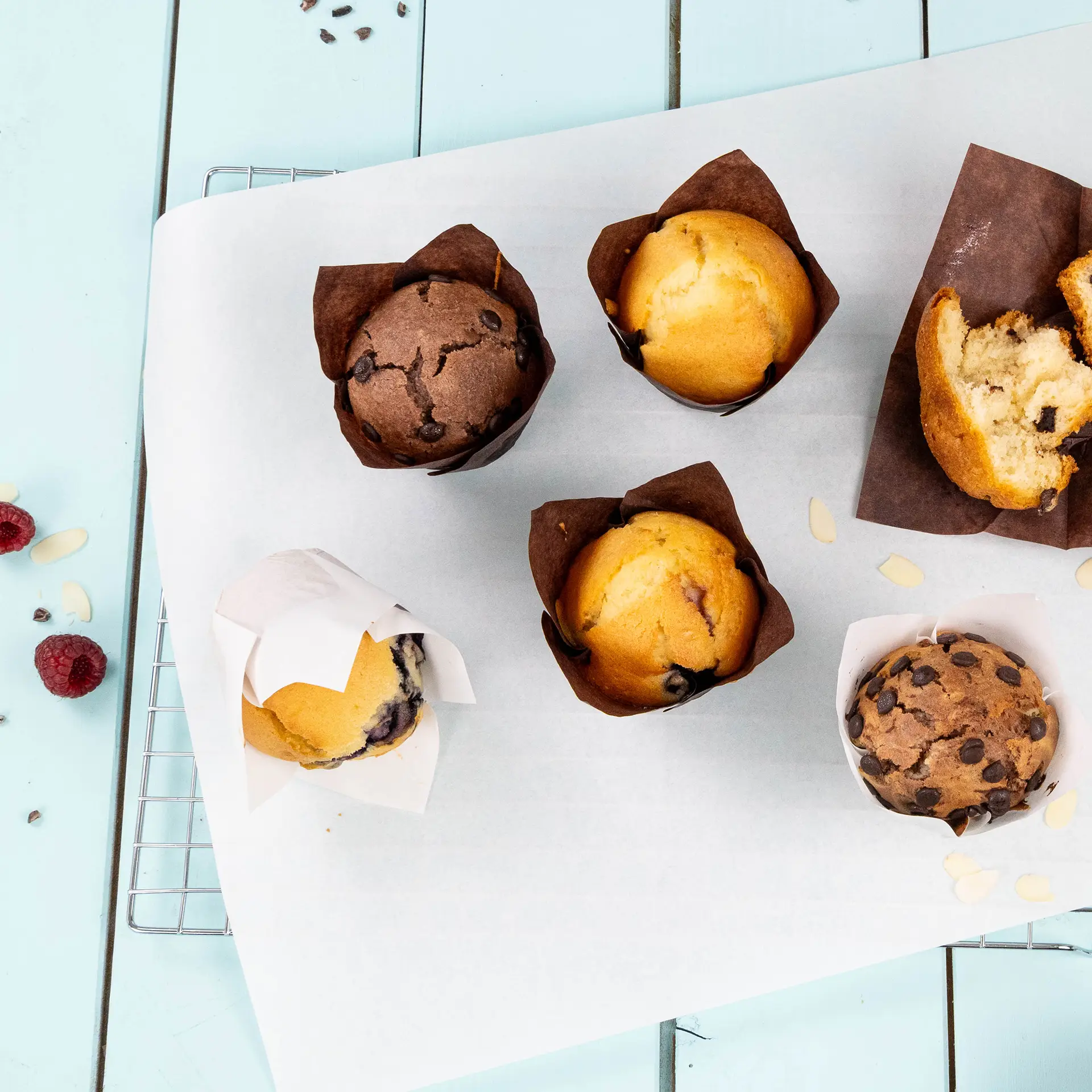 Muffins variés dans des gobelets en papier imperméable marron sur une plaque de cuisson blanche, avec des framboises et des éclats d'amande à proximité.