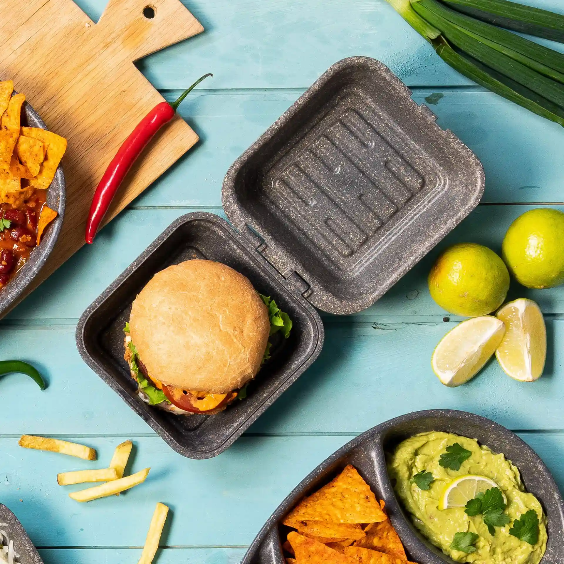 Boîte à burger grise réutilisable avec couvercle, contenant un burger, entouré de citron vert, de nachos et d'un piment.