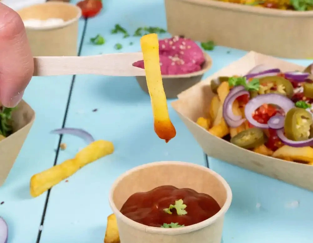 Une main tient un bâton en bois avec une frite au-dessus d'un verre de ketchup posé sur une surface bleue.