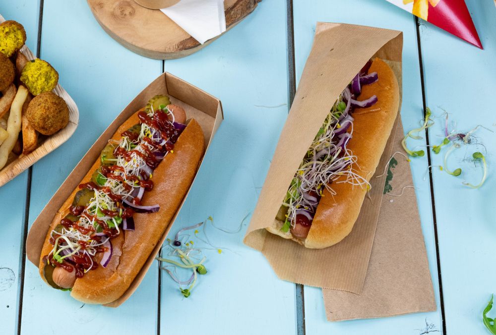 Deux hot-dogs avec divers garnitures, y compris des cornichons, des oignons et des germes, servis dans des contenants en papier sur une table bleue.