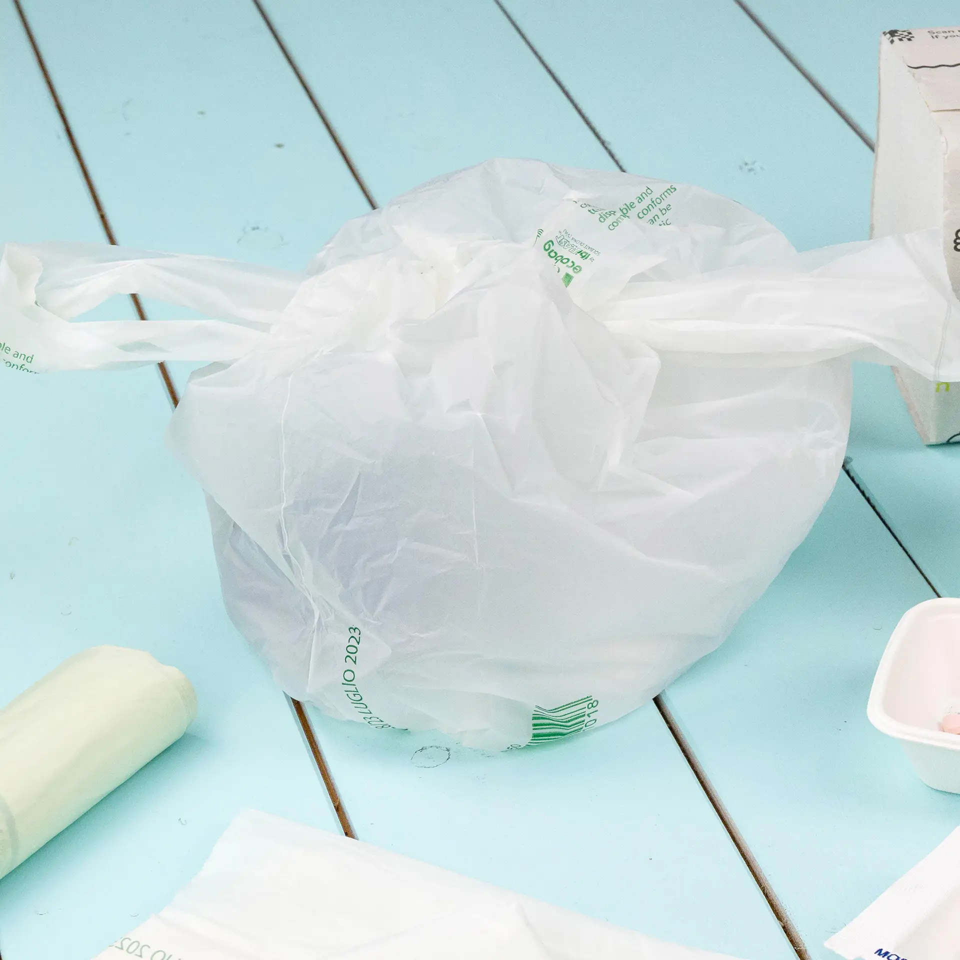 Un sac poubelle transparent en bioplastique, en PLA, noué en haut ; d'autres éléments éparpillés à proximité sur une surface bleue.