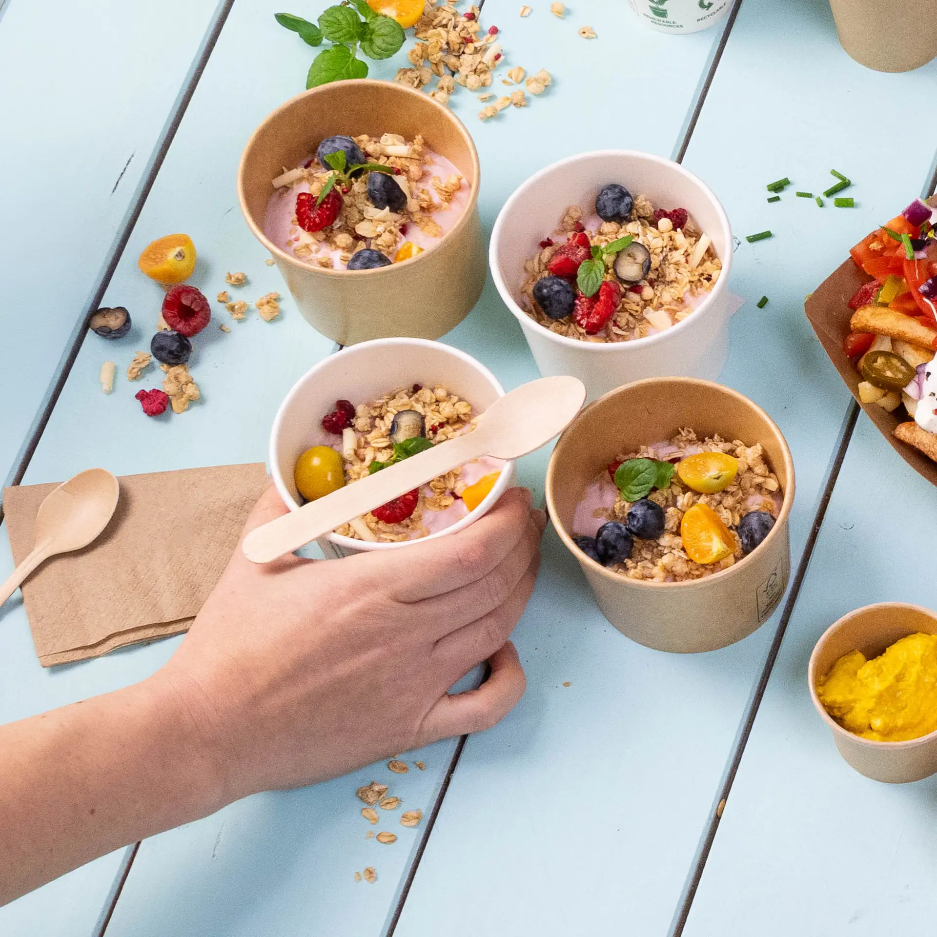 Une main tenant une petite cuillère en bois au-dessus de plusieurs pots de yaourt garnis de granola et de fruits sur une table bleue.
