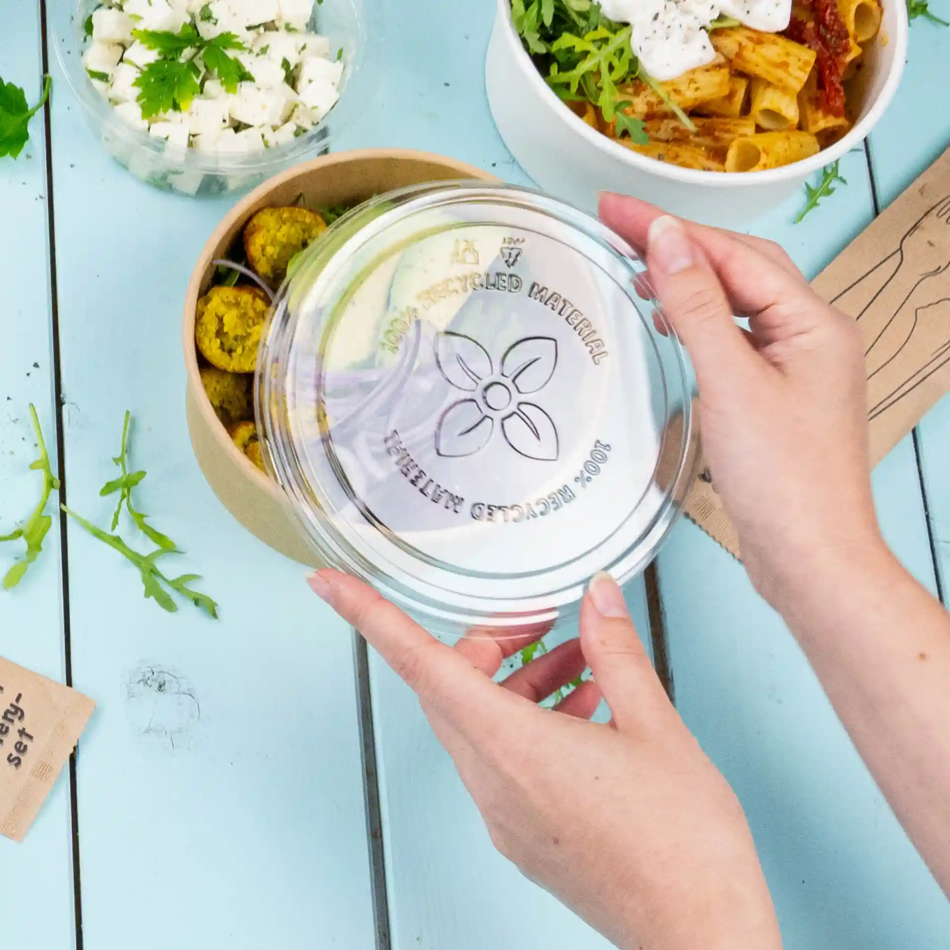 Des mains tenant un couvercle en rPET transparent au-dessus d'un récipient rond avec des falafels, sur une surface bleue avec des aliments à proximité.