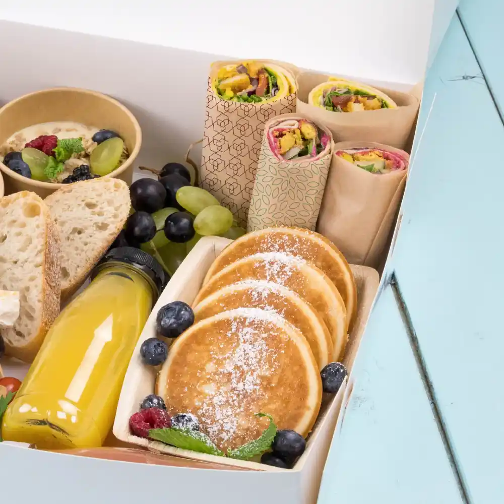 Une variété d'aliments comprenant des wraps, des crêpes, des fruits et une bouteille de jus dans une boîte à emporter sur une surface bleue.