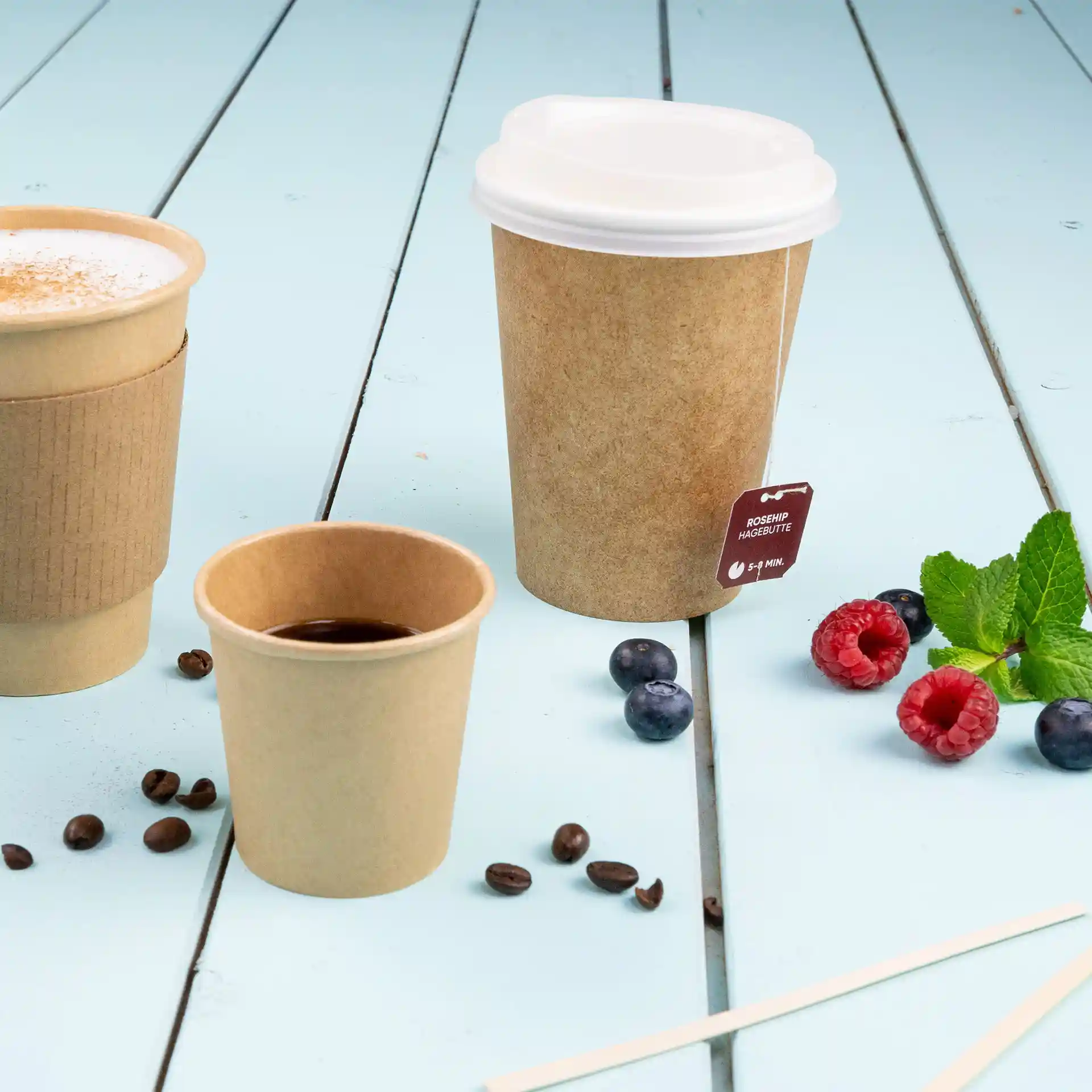 Image de trois gobelets en papier sur une surface en bois bleu ; l'un est un gobelet marron de 100 ml avec un couvercle blanc, un autre est un gobelet marron plus petit, et le troisième est rempli de café.