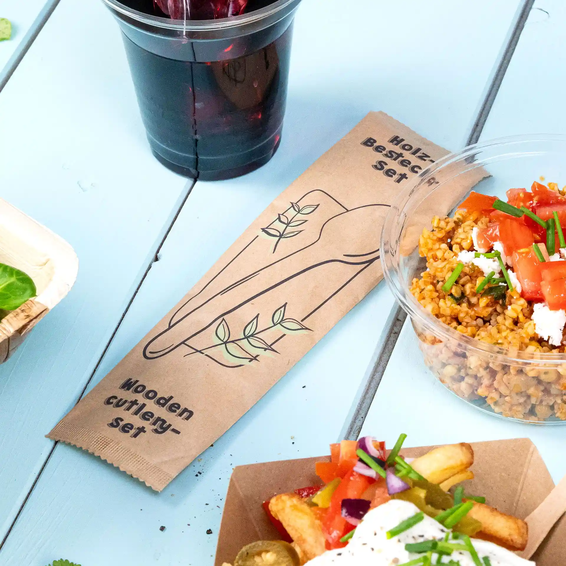 Emballage d'un ensemble de couverts en bois avec un design d'ustensiles et de feuilles, placé à côté d'aliments sur une surface bleue.