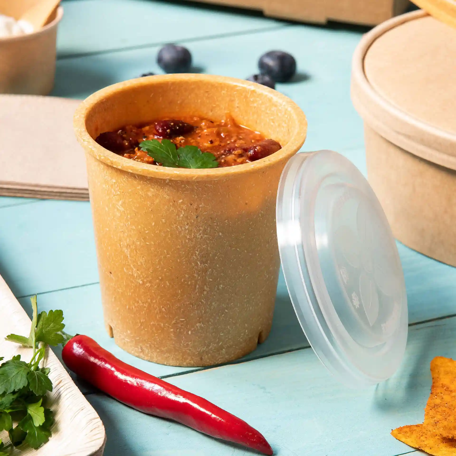 Gobelet réutilisable marron avec couvercle, rempli de nourriture, posé sur une surface bleue avec un piment rouge et des herbes à proximité.
