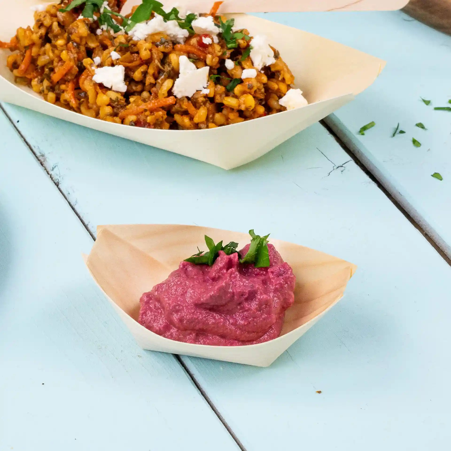 Plat en forme de bateau en bois avec du houmous rose garni d'herbes hachées, posé sur une surface bleu clair.