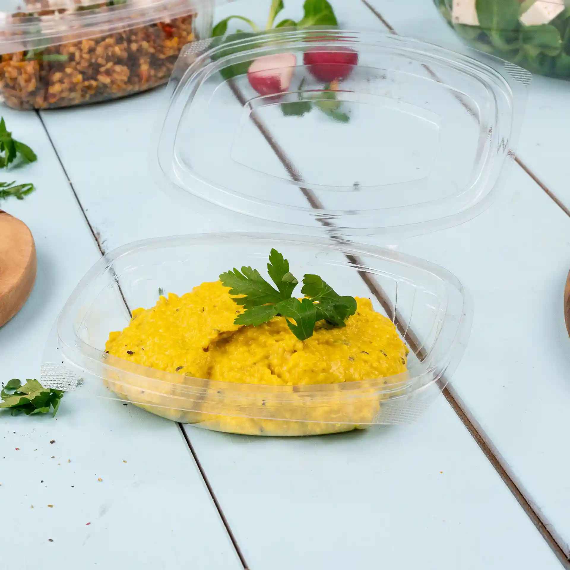 Bol transparent rectangulaire avec un couvercle pliant, contenant des aliments jaunes surmontés d'une branche de persil.