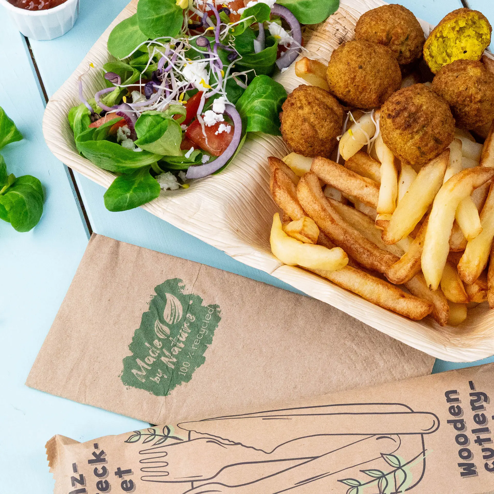 Une assiette avec des falafels, des frites et une salade mixte ; emballage en couverts en bois visible ; serviette en papier étiquetée 'Made by Nature.'