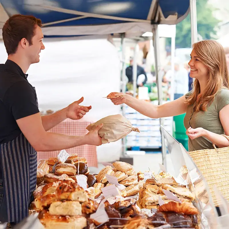Un vendeur remet un sac en papier brun à un client dans un stand de marché présentant diverses pâtisseries.