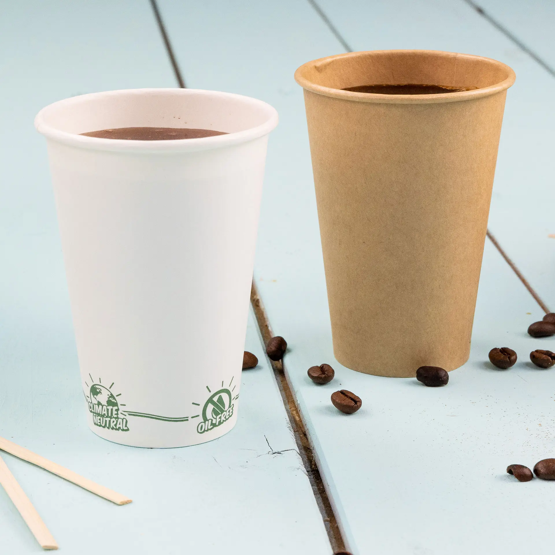 Un gobelet en papier blanc avec un logo vert, à côté d'un gobelet en carton marron ; des grains de café éparpillés sur une table bleue.