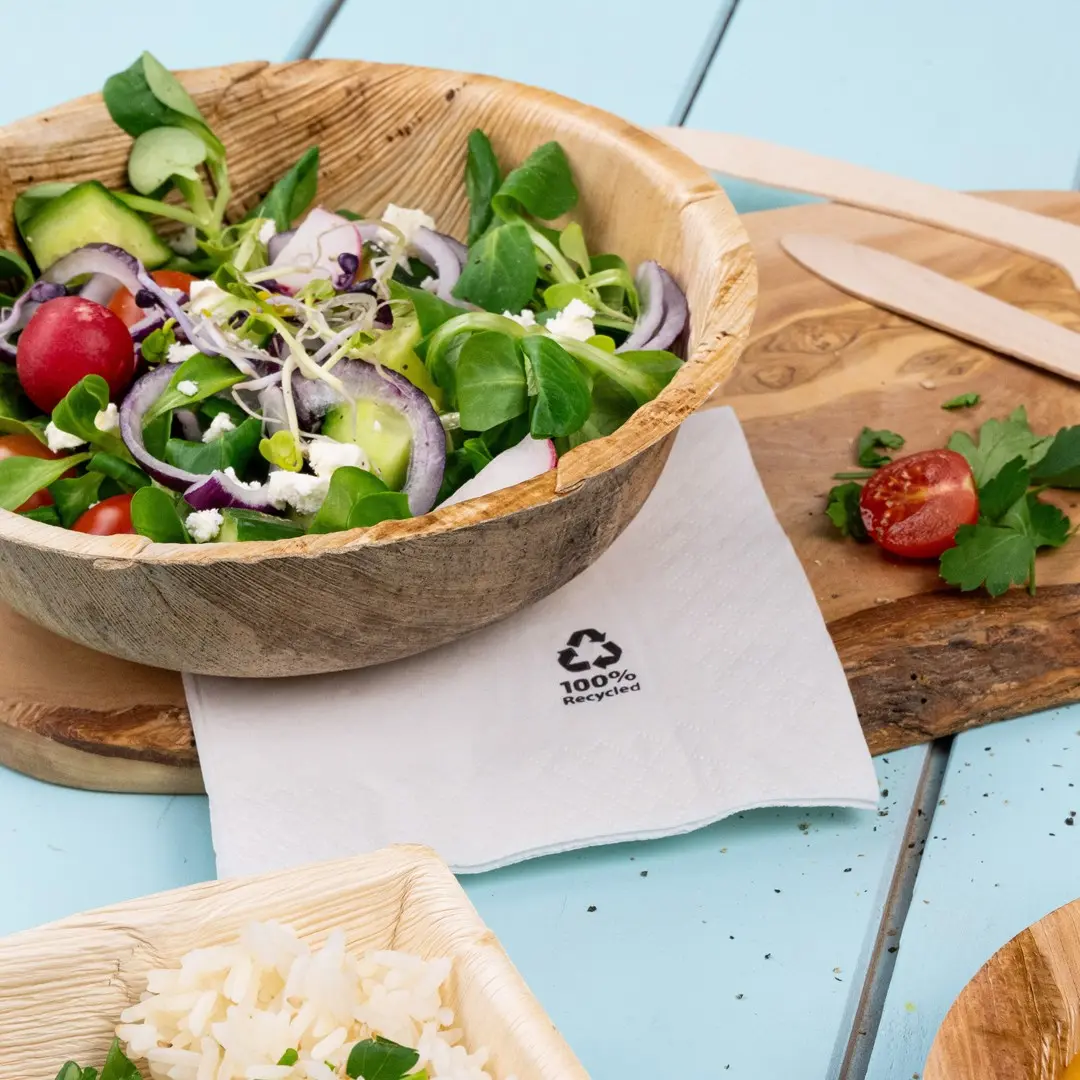 Salade dans un bol en bois avec une serviette étiquetée "100% Recycled", à côté de riz et d'une tomate cerise sur une planche en bois.