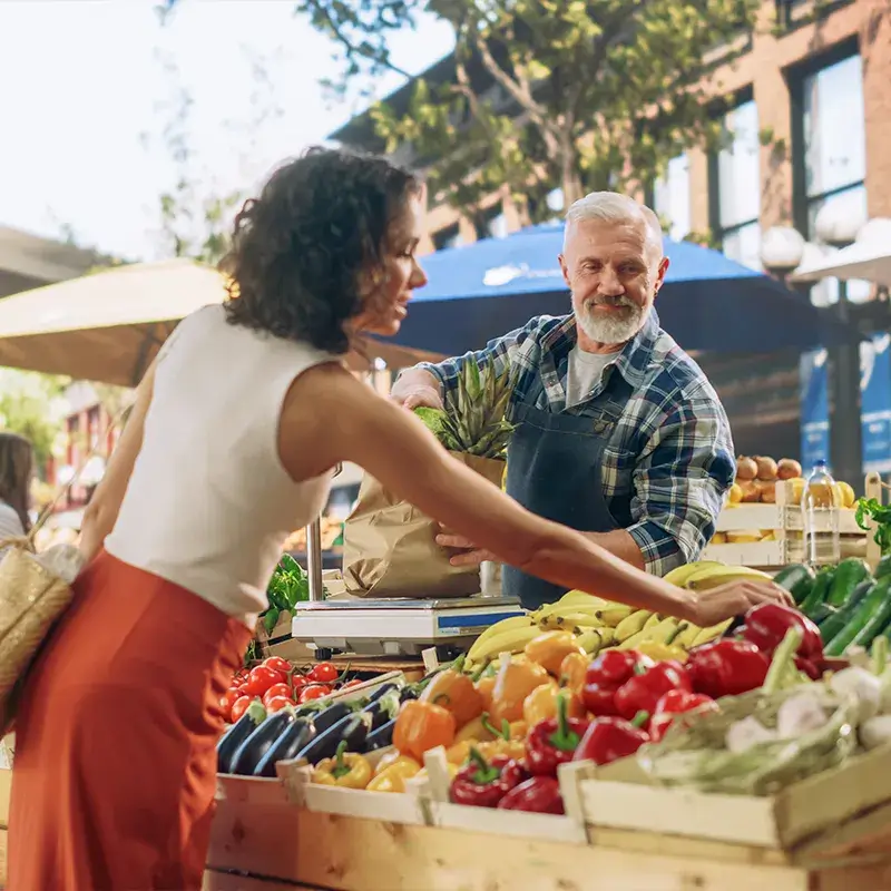 Une femme en jupe rouge achète des légumes auprès d'un vendeur à un stand de marché rempli de produits frais.